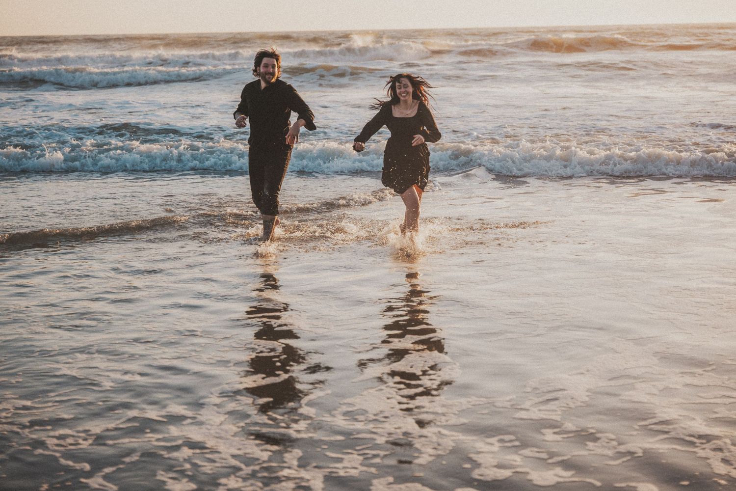 Couple running joyfully through ocean waves on a sandy beach at golden hour sunset.