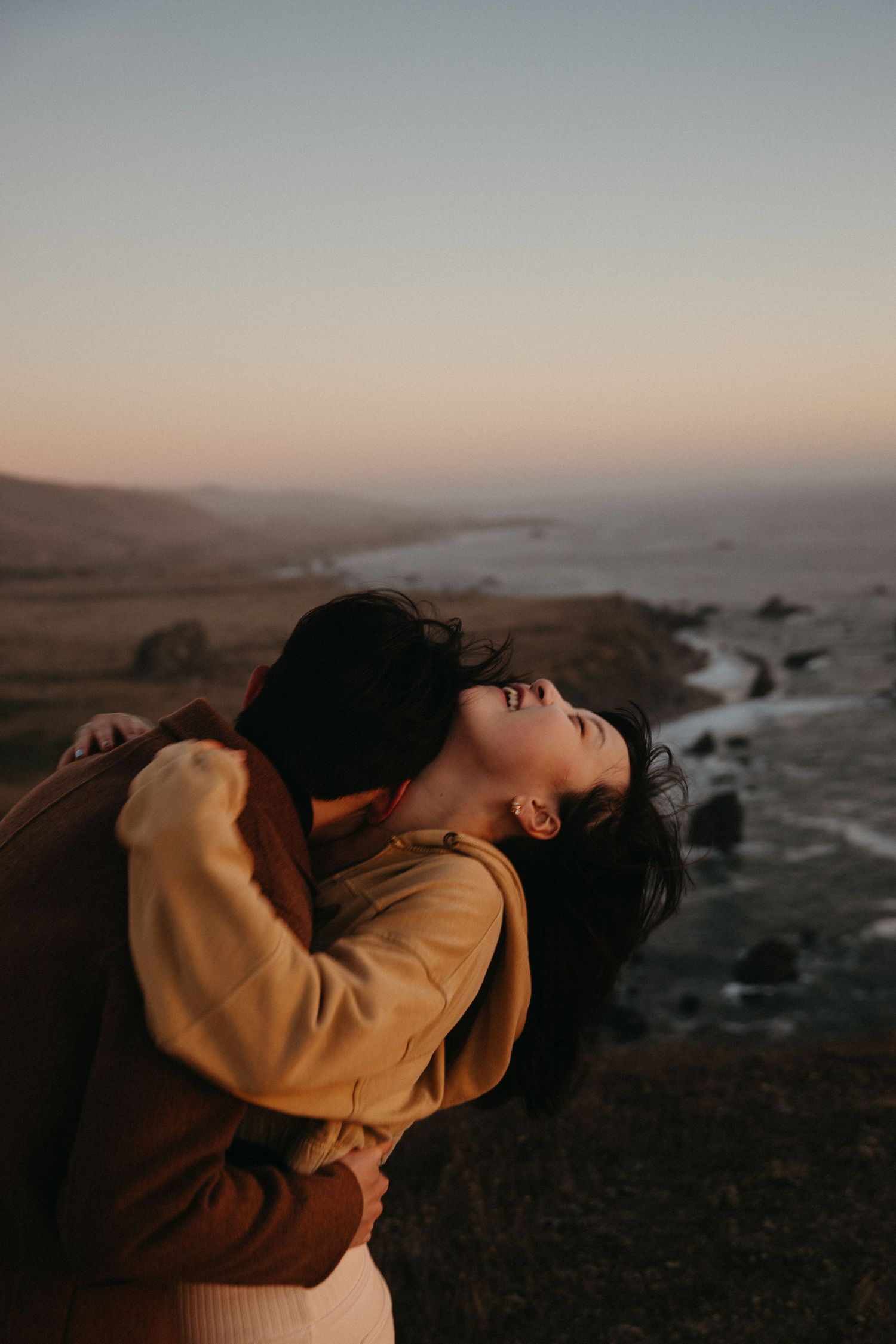 A couple embraces on a rugged coastline at sunset as the woman tilts her head back toward the soft pastel sky.