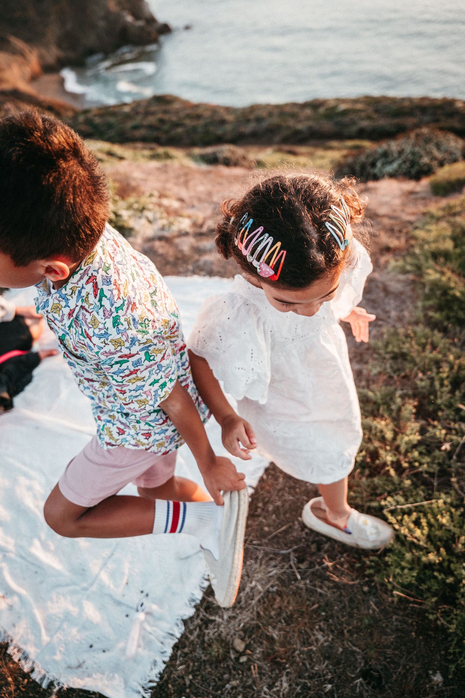 Two children setting up a white picnic blanket on a coastal cliff overlooking the ocean at sunset.