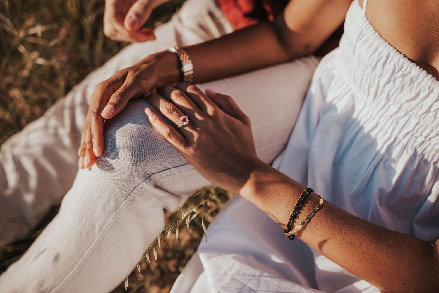 Close-up of a couple holding hands outdoors, woman wearing multiple rings and bracelets, sitting together in a field.