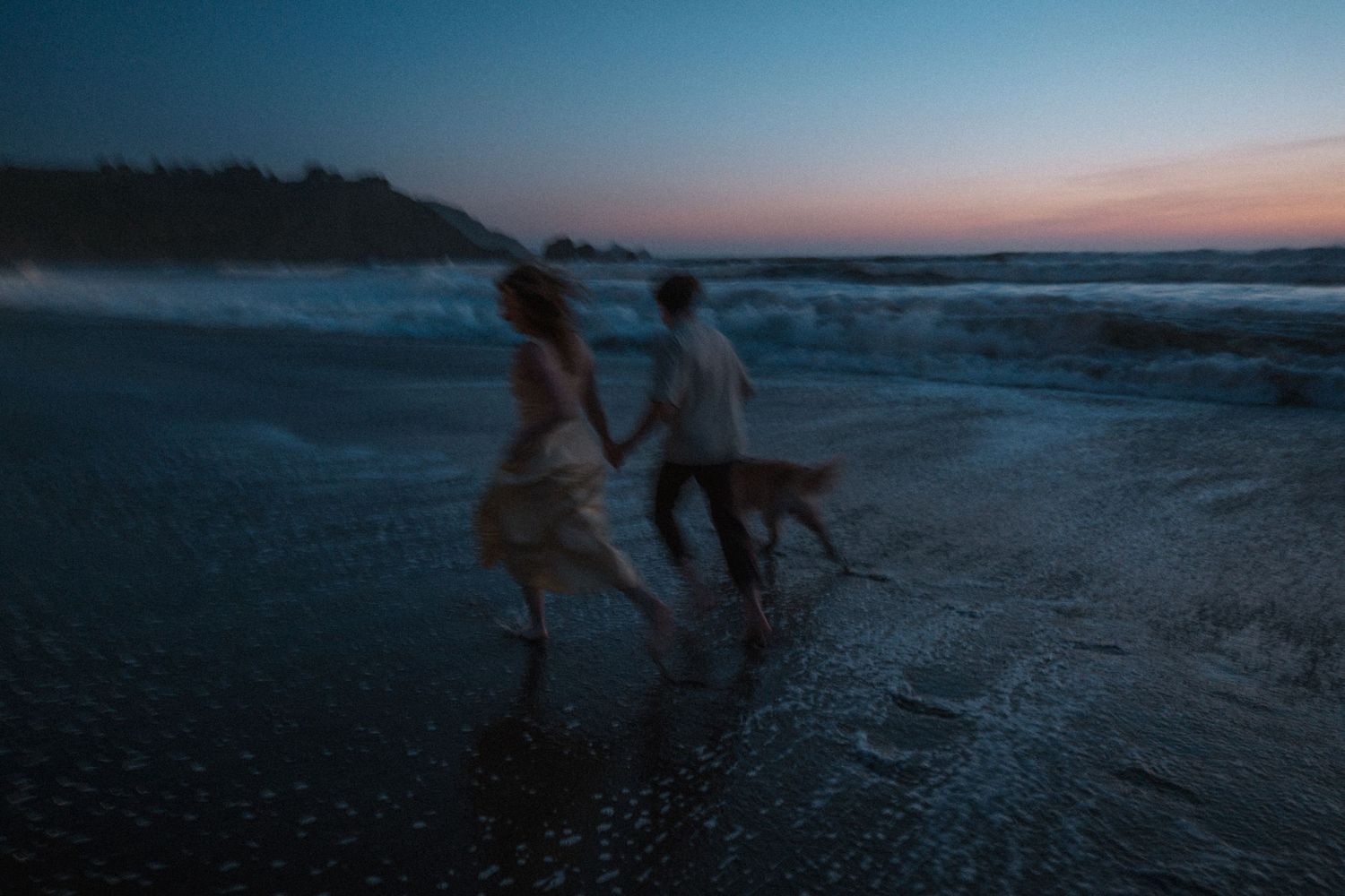Blurred motion shot of couple running on beach at dusk with dog in crashing waves.