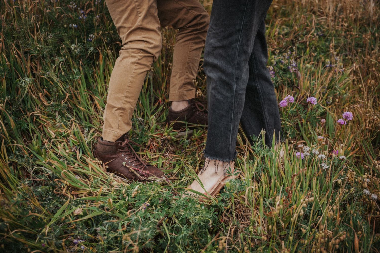 Two people standing close together in a wildflower meadow, wearing casual pants and boots on a romantic outdoor date.