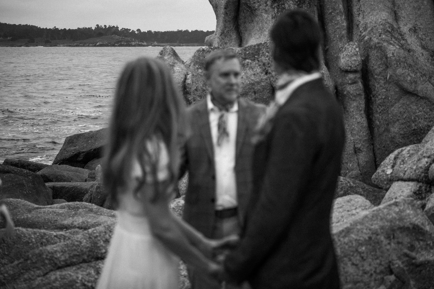 Black and white photo of a couple holding hands during an outdoor wedding ceremony by rocky ocean shore.Photography by The Y.K. Studio.