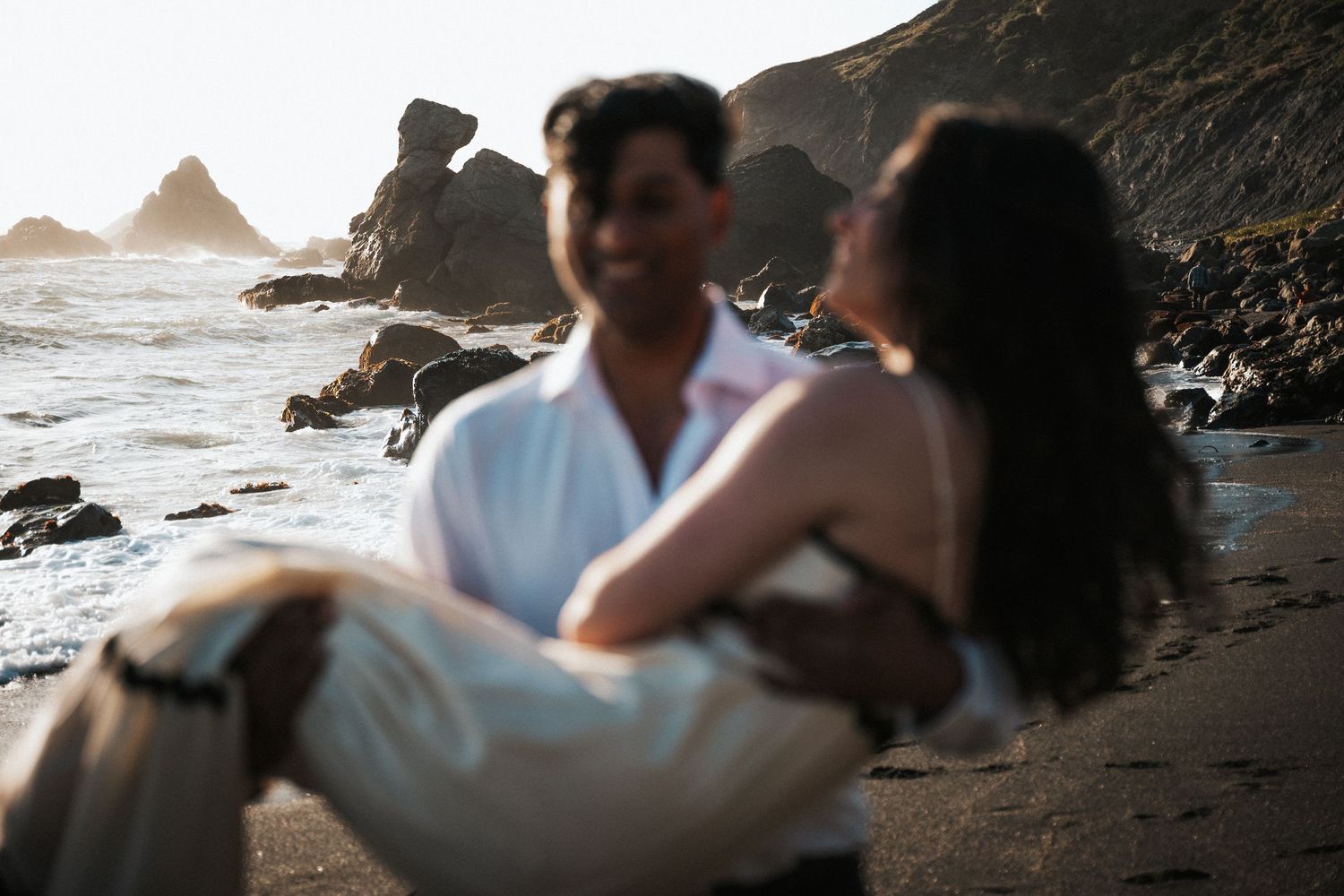 Couple sitting together on a rocky beach at sunset, sharing a romantic embrace by the ocean waves.