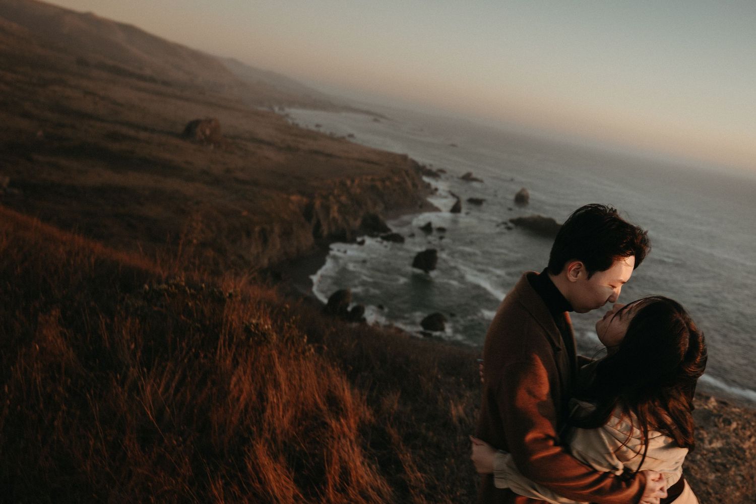 Couple sharing an intimate kiss on a rugged clifftop at dusk, with waves crashing on rocks and warm sunset light over the coast.