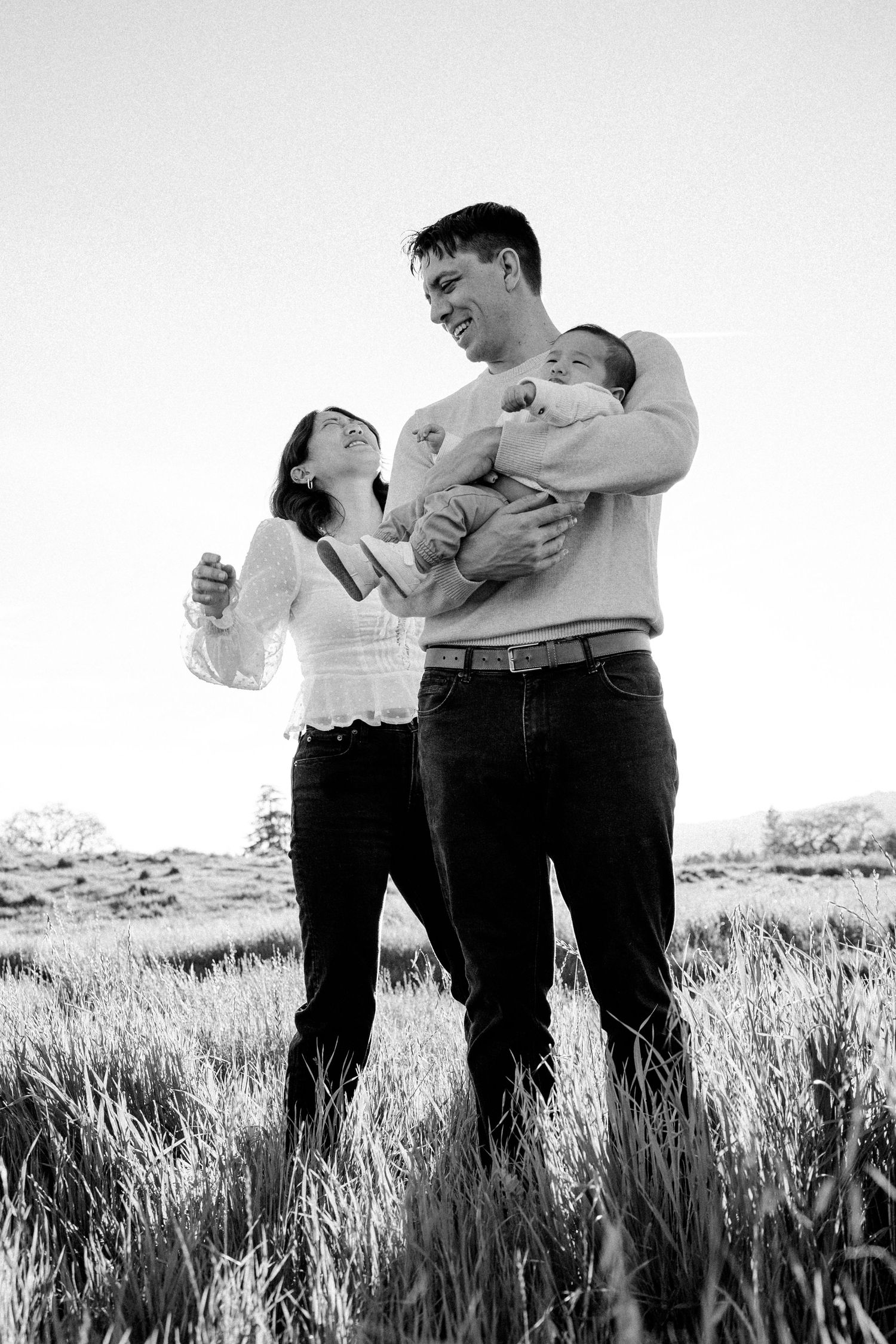 Happy couple holding newborn baby in sunlit grass field black and white family portrait.