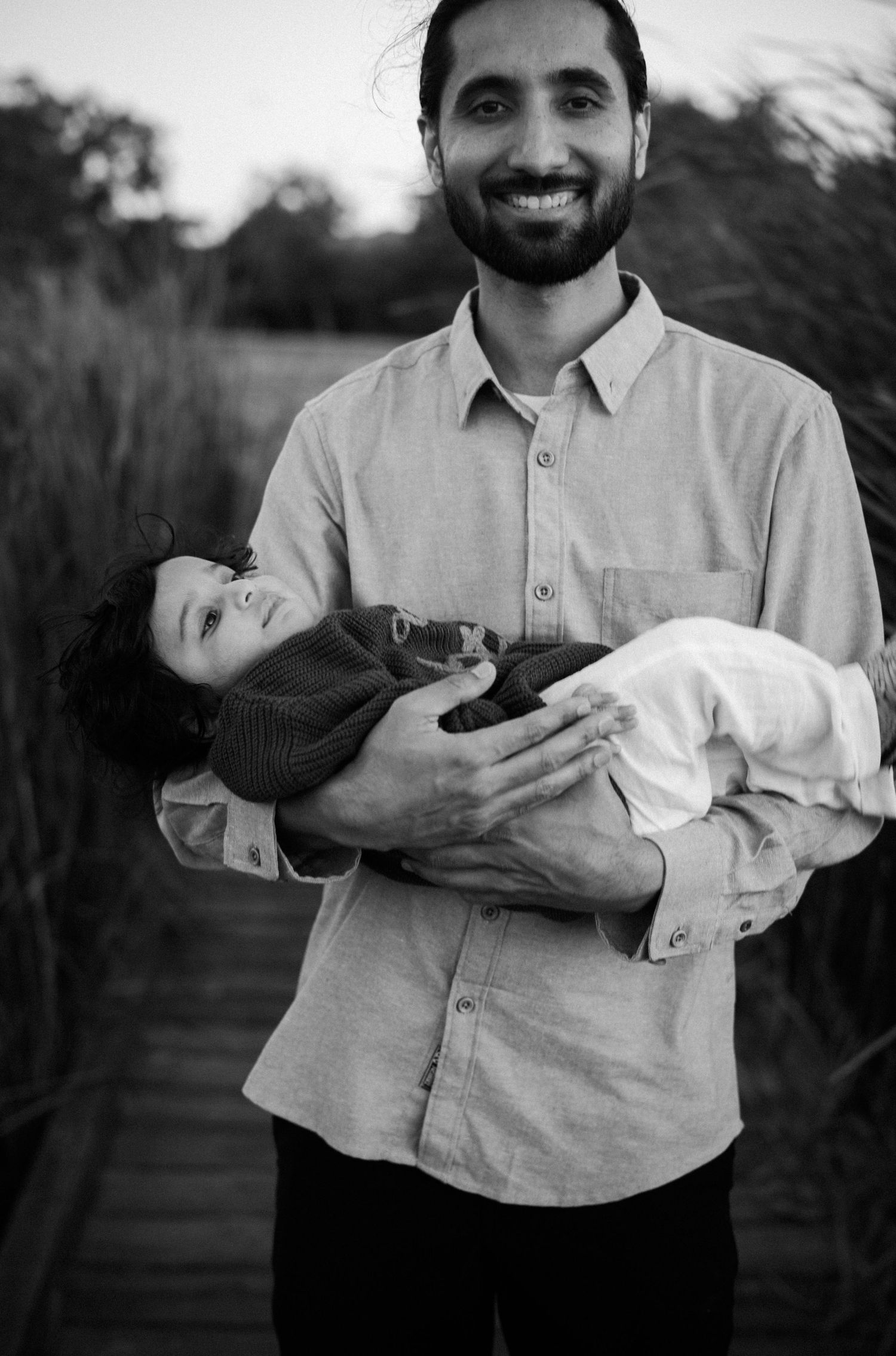 Smiling bearded father holds sleeping baby in black and white outdoor portrait.