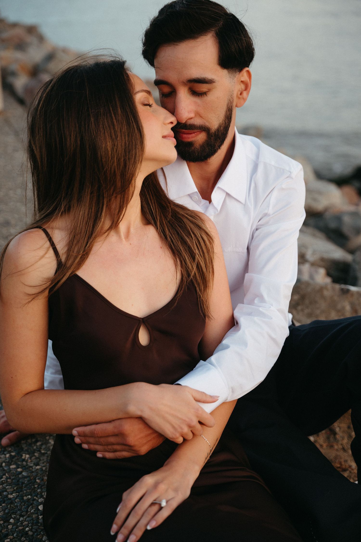 Romantic couple sharing a tender moment by the ocean, woman in brown dress with engagement ring, man in white shirt.