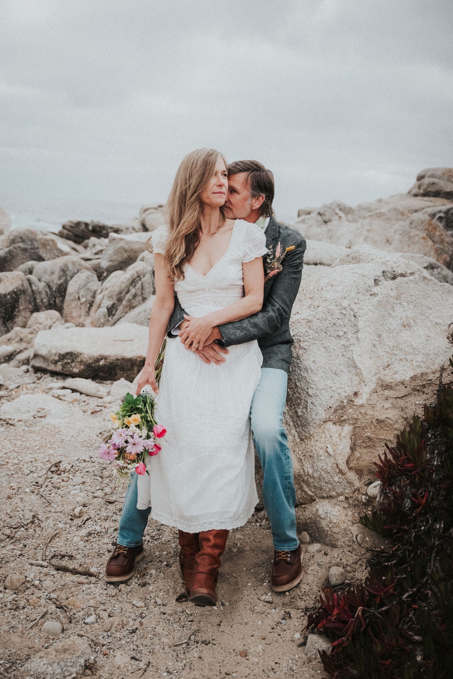 Couple embraces on rocky coastal shore, bride in white dress holding bouquet, groom kissing her cheek.