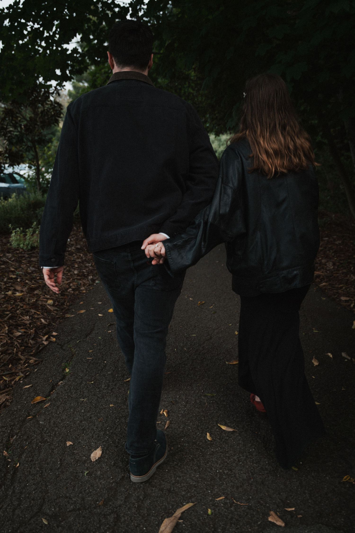 Couple holding hands walking on a path surrounded by autumn leaves and trees, both dressed in black outfits.
