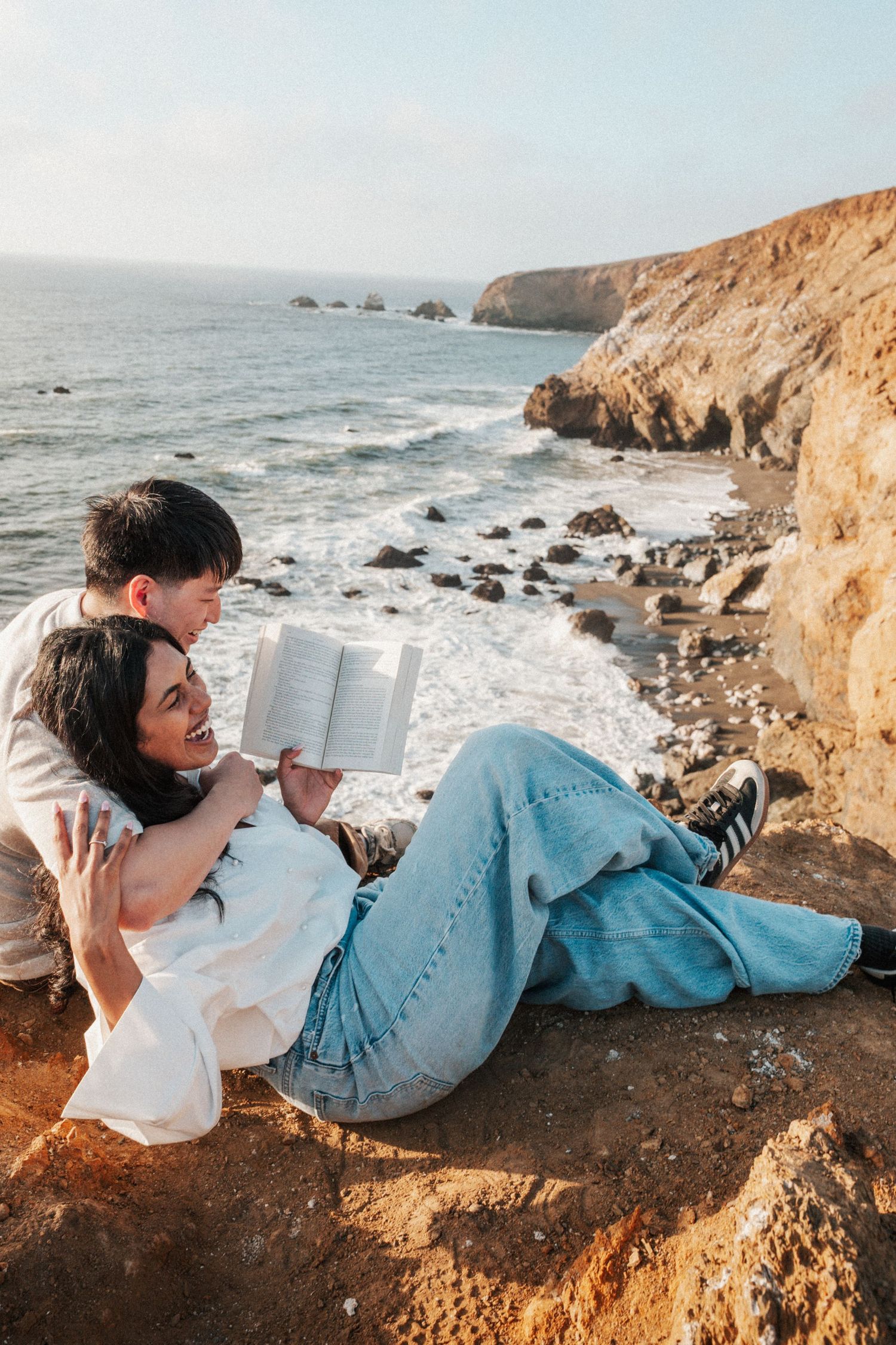 Couple relaxing on coastal cliffs, woman reading book while leaning against partner with ocean waves in background.