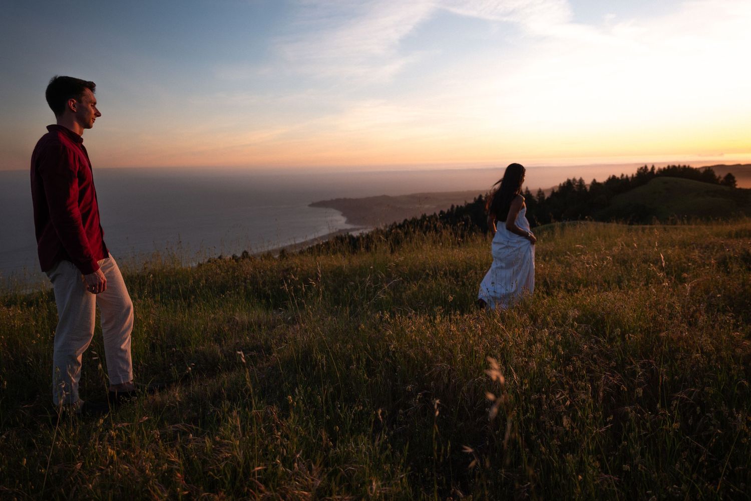 Couple standing apart in wildflower meadow overlooking coastal cliffs and ocean during vibrant golden sunset.