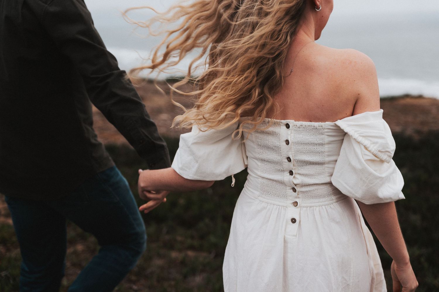 Couple holding hands outdoors, woman in white off-shoulder dress with buttons, hair blowing in wind.