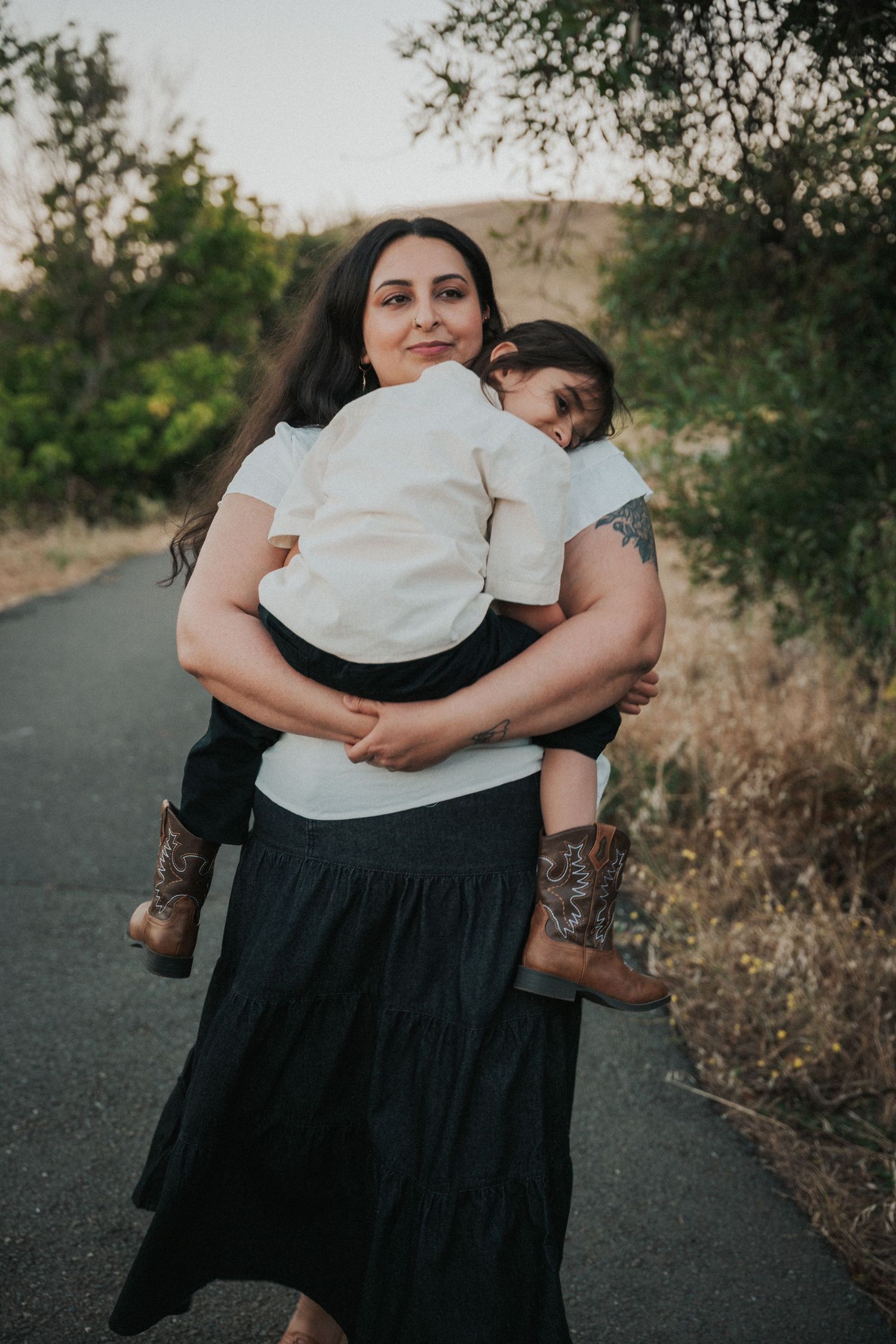 A mother tenderly holds her child on a scenic path, both wearing white tops, with green trees in the background.