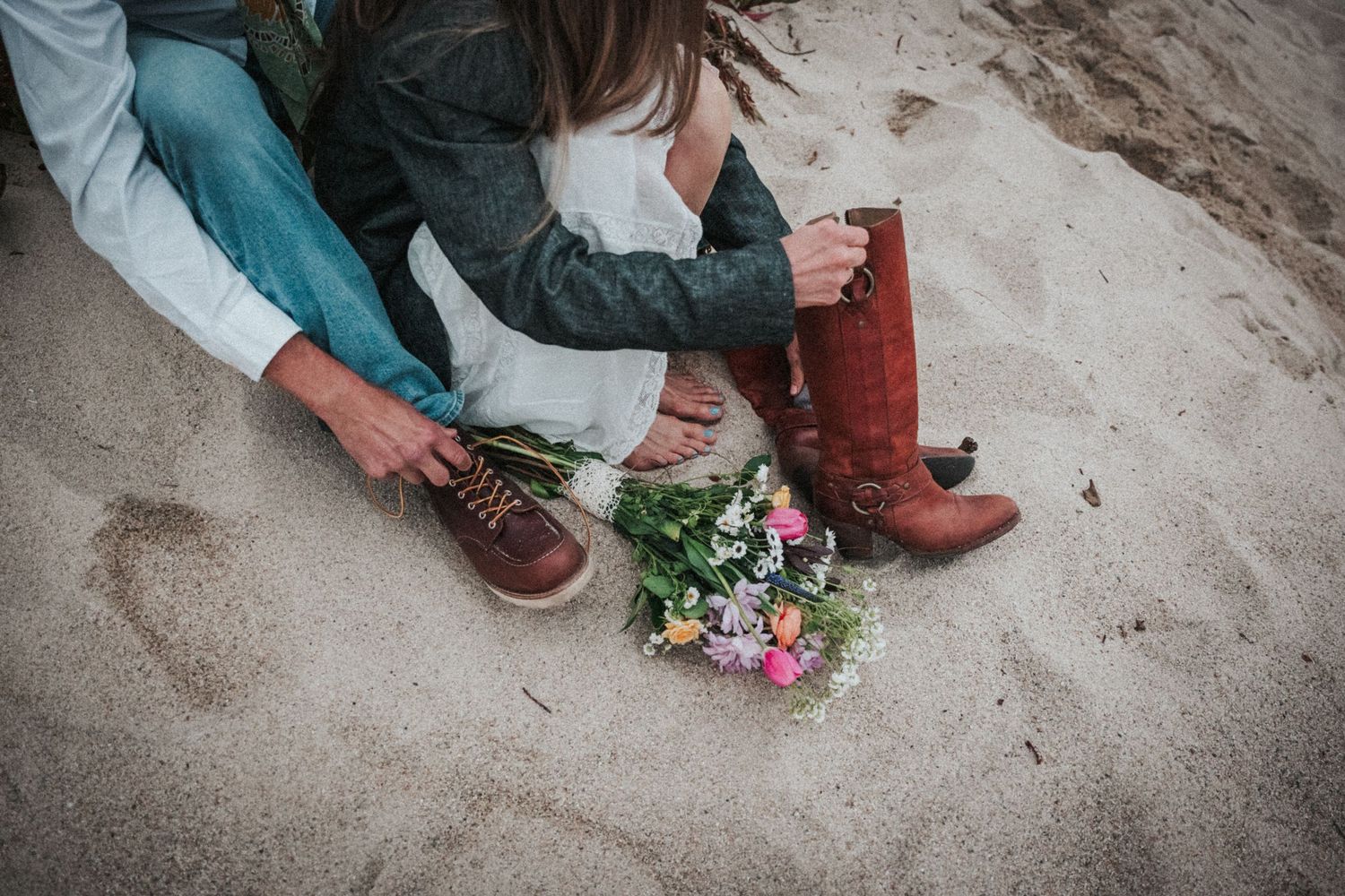 A couple sits on sandy beach with a colorful wildflower bouquet between them.
