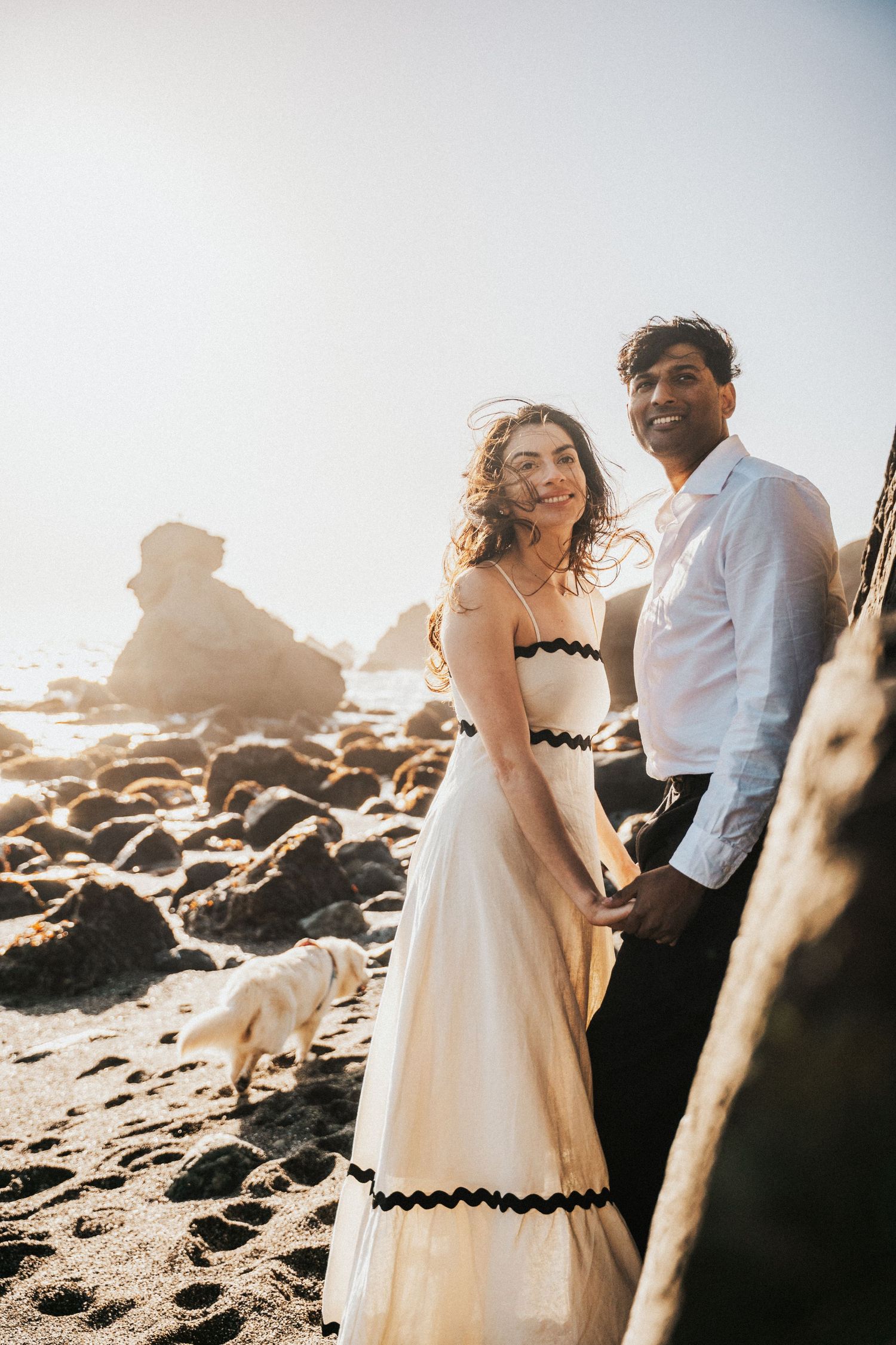 Couple holding hands on a rocky beach at golden hour, woman in white dress with black trim, man in white shirt, dog nearby.