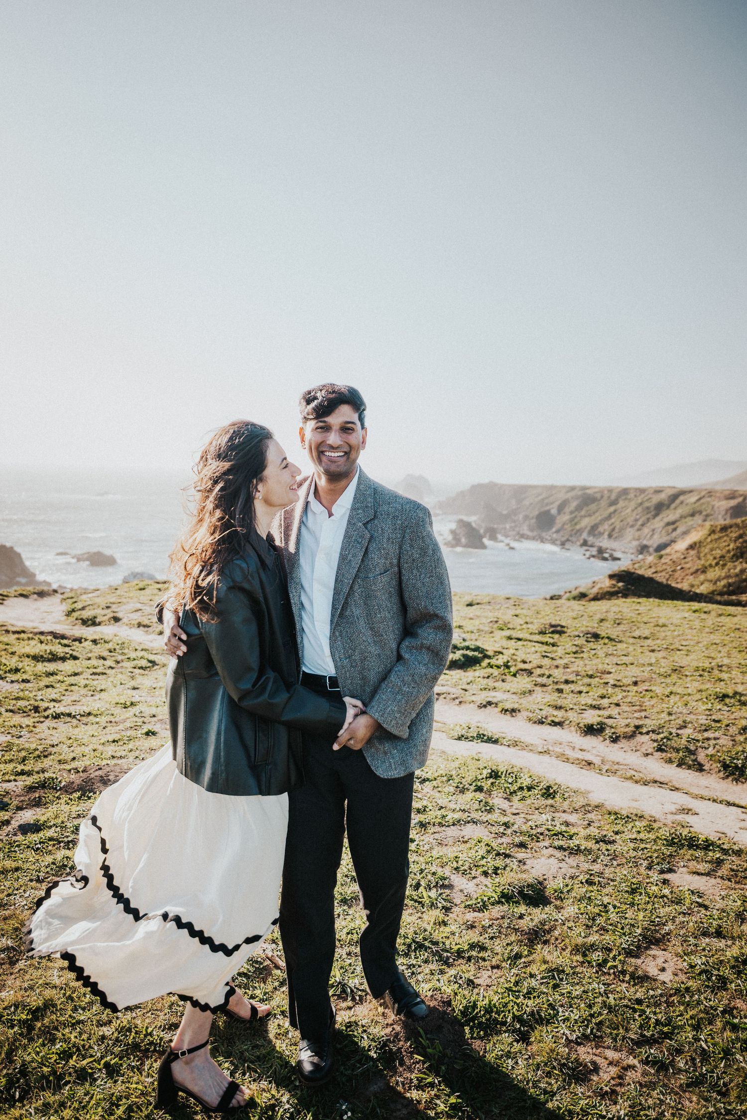 Couple embracing on coastal cliffside, woman in white skirt and leather jacket, man in grey blazer, ocean backdrop.