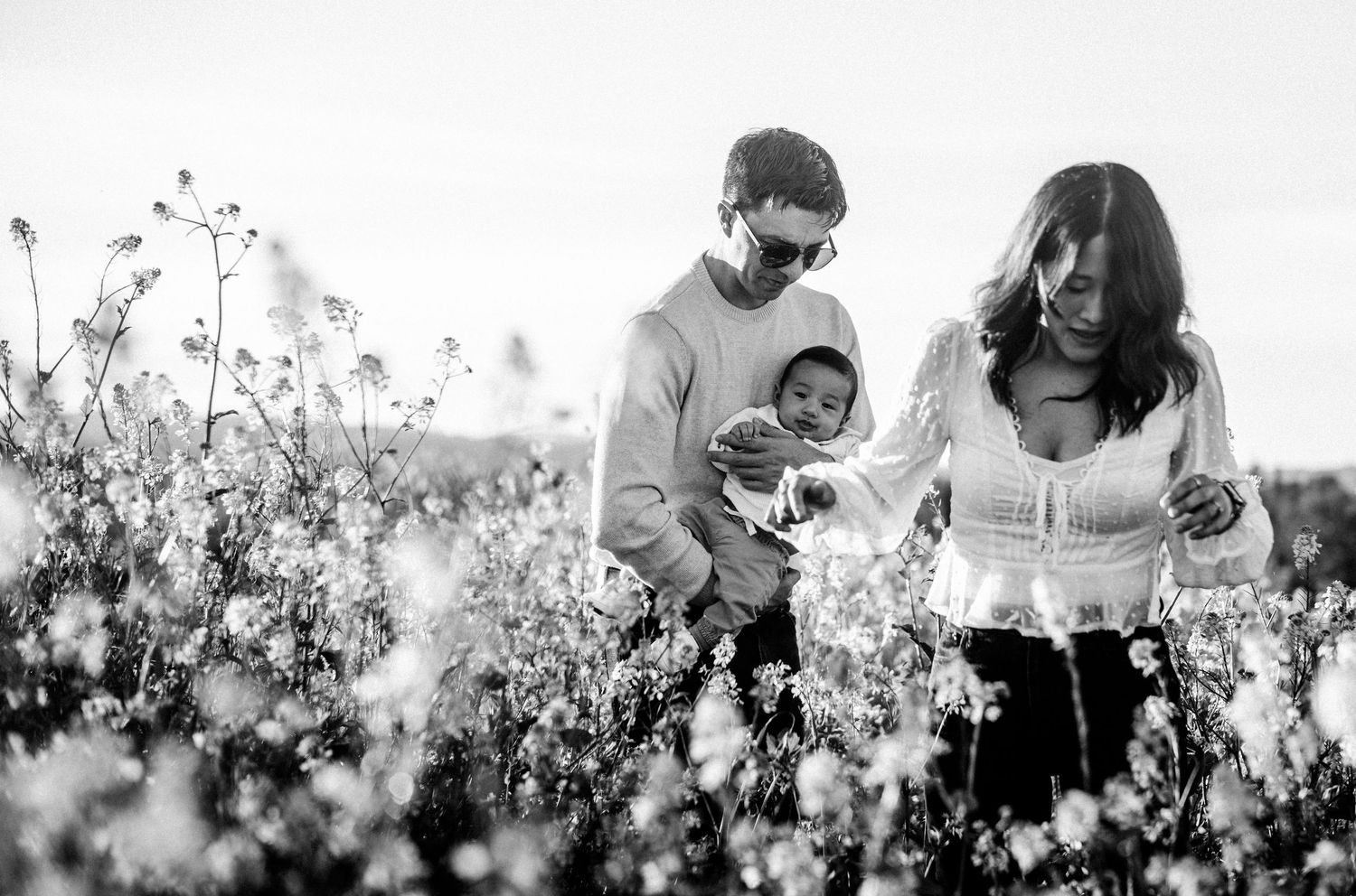 Black and white photo of a couple holding their newborn baby in a blooming wildflower field.