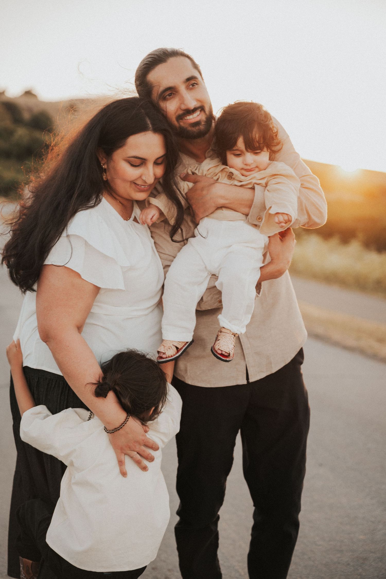 Family of four posing outdoors at golden hour, parents smiling warmly while holding young children in a heartfelt moment.