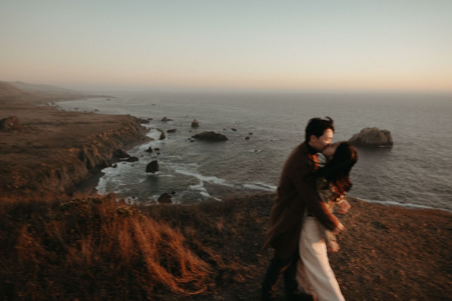 Couple embracing on a windswept seaside bluff at golden hour with rocky coastline and calm ocean behind them.