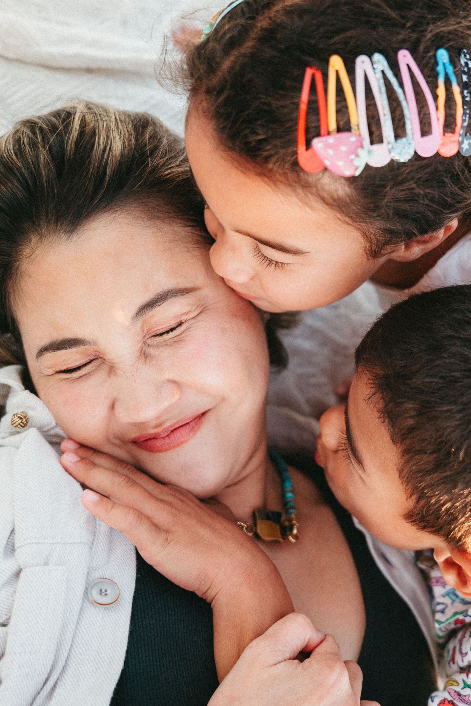 A woman smiles warmly as two children cuddle and kiss her cheeks in a sweet family moment.