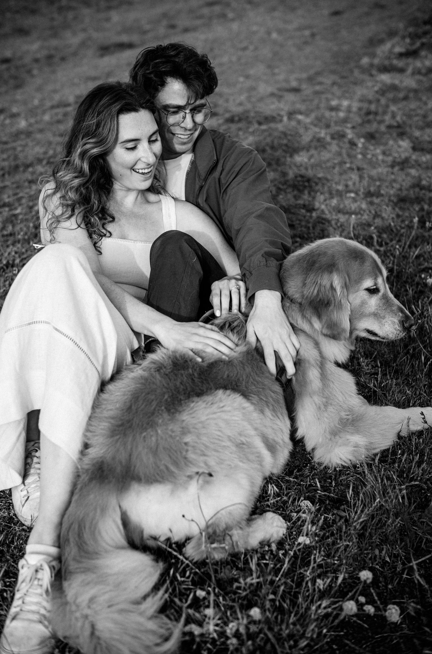 Black and white photo of smiling couple sitting in grass with golden retriever dog.