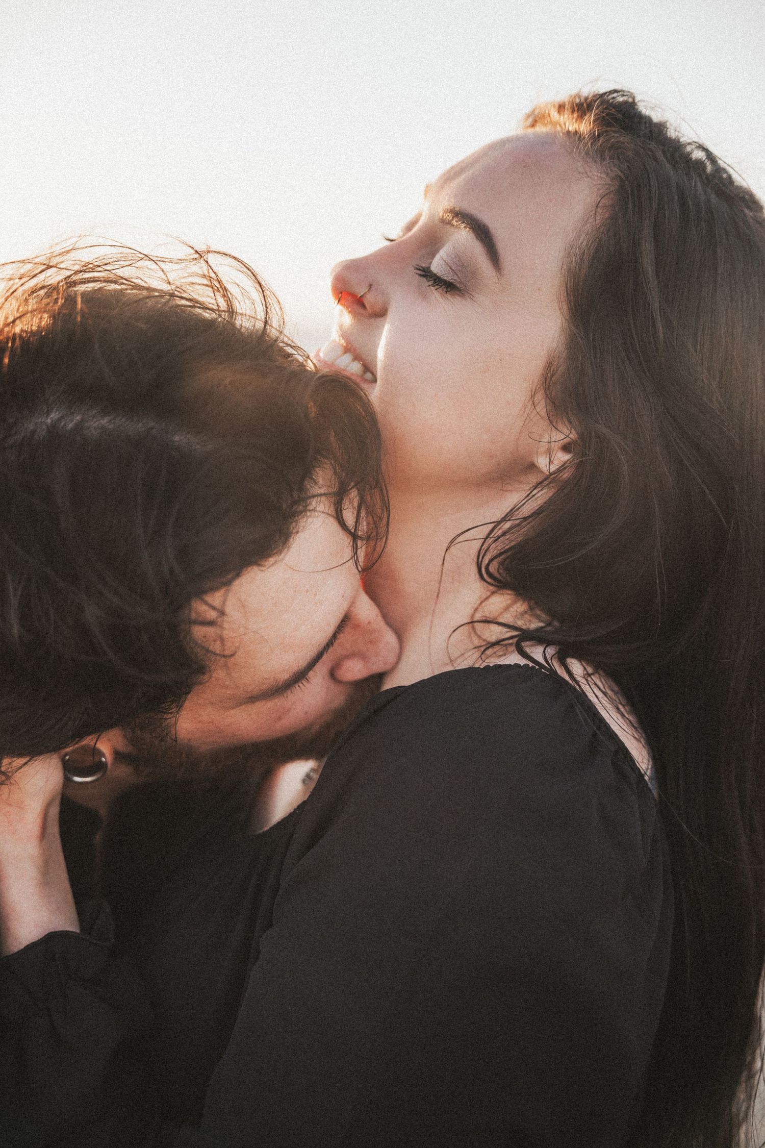 Couple sharing an intimate moment outdoors, man kissing woman's neck in warm backlit sunlight. Photography by The Y.K. Studio.