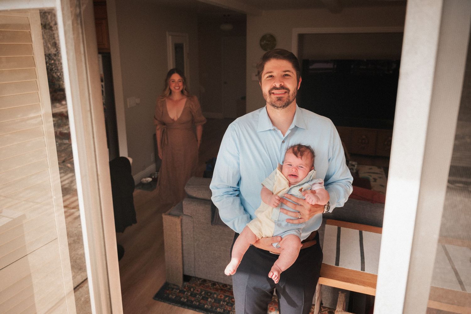 A smiling man holds a baby near a doorway while a woman stands in the background inside a cozy home.
