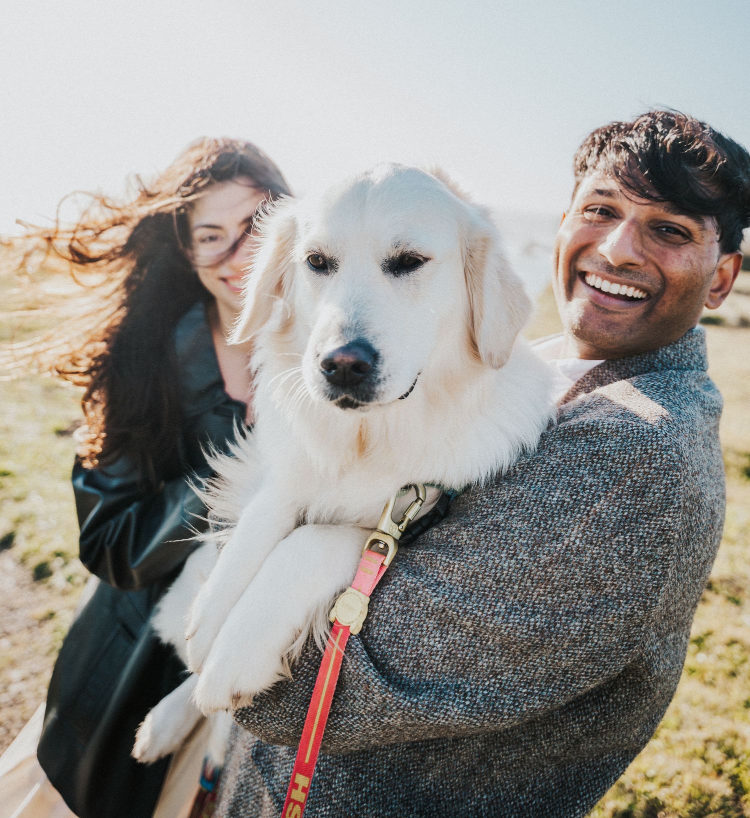 A smiling man holds a large white dog with a red leash while a woman with curly hair stands beside him outdoors.