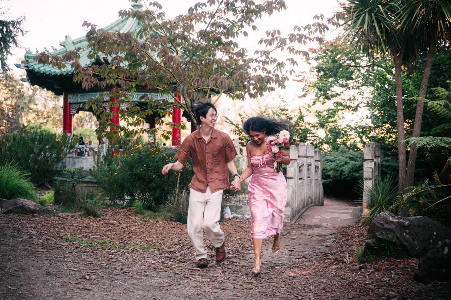 Couple holding hands and running through a scenic Chinese garden with a pagoda and lush greenery.