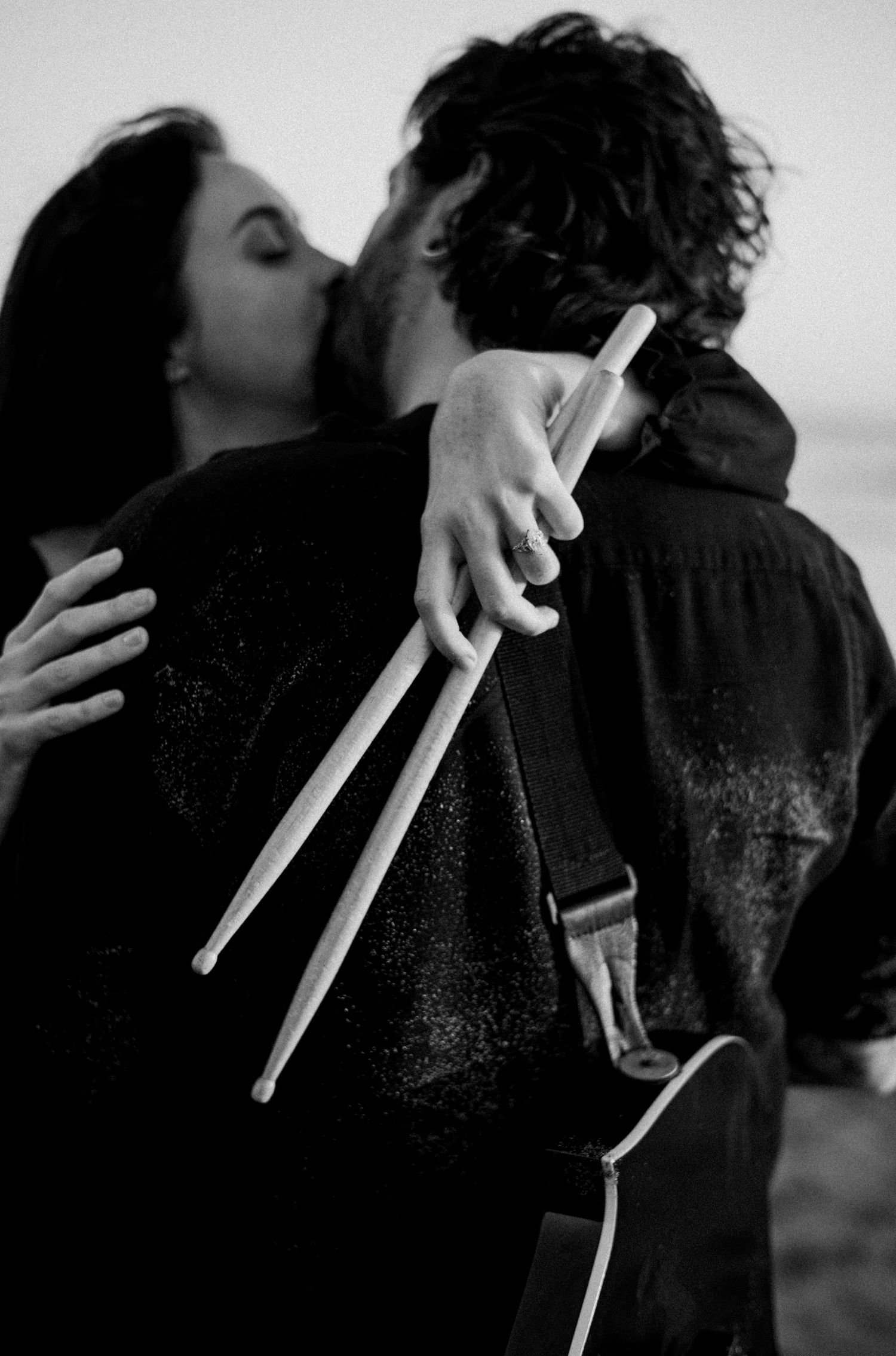 Black and white photo of a couple kissing while embracing, one holding drumsticks outdoors.