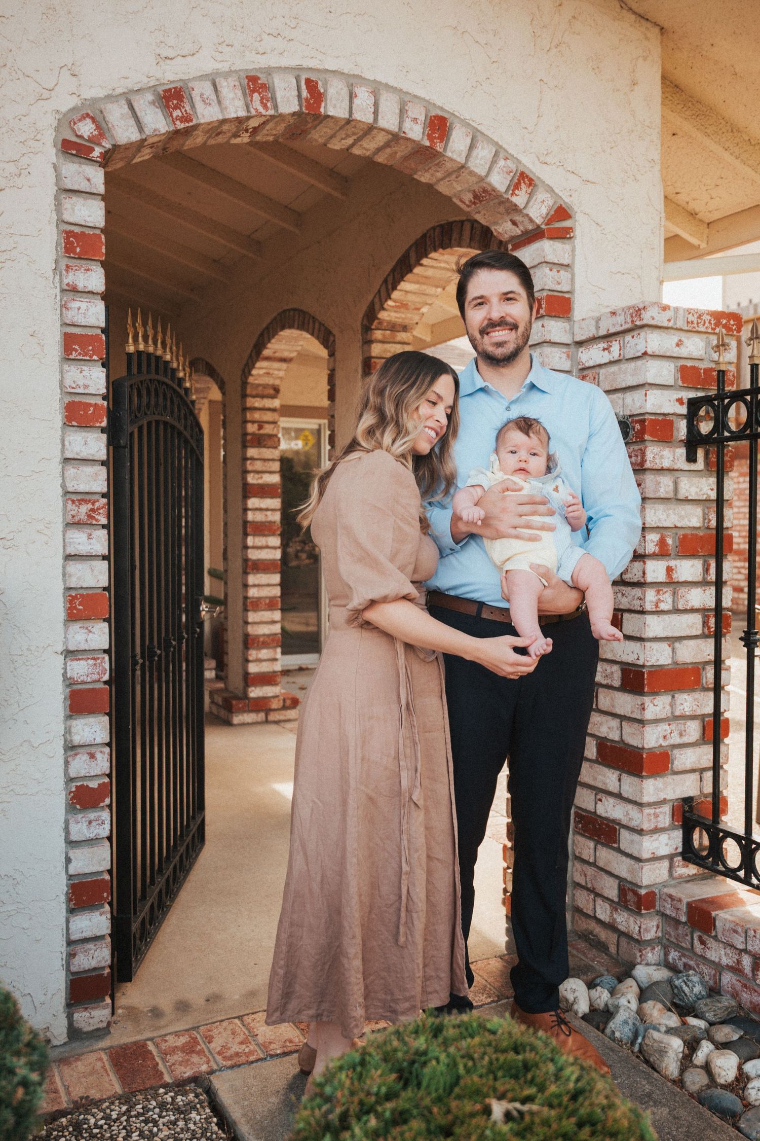 Young family with baby posing at brick archway entrance of home, mother in mauve dress, father in blue shirt.
