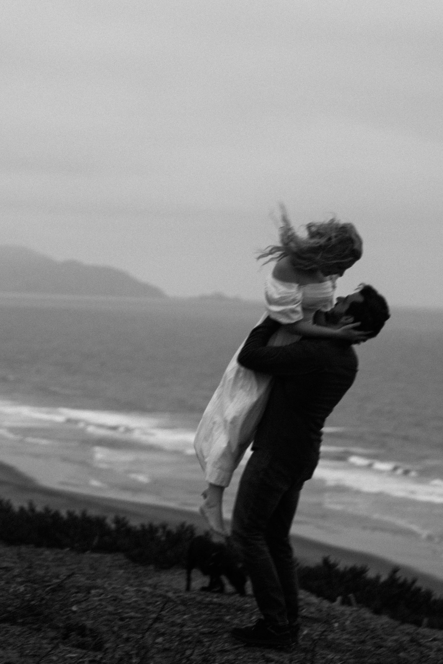 A couple shares a romantic embrace on a windy beach, the man lifting the woman in a white dress against an overcast sky.