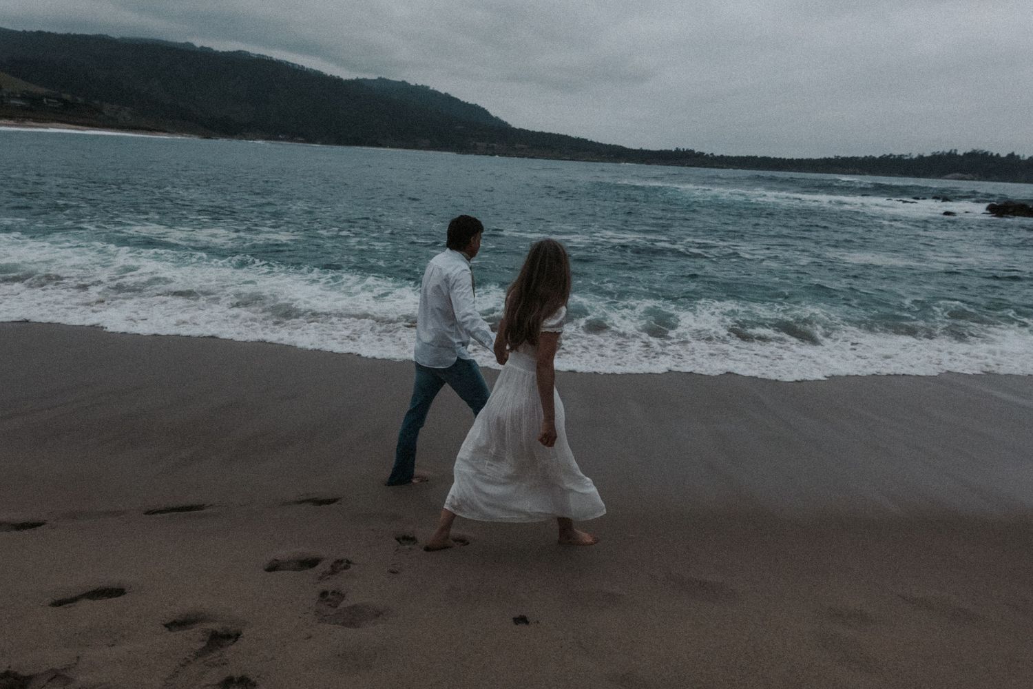 Couple walking hand in hand on a sandy beach with ocean waves, wearing white dress and casual attire on a cloudy day.Photography by The Y.K.