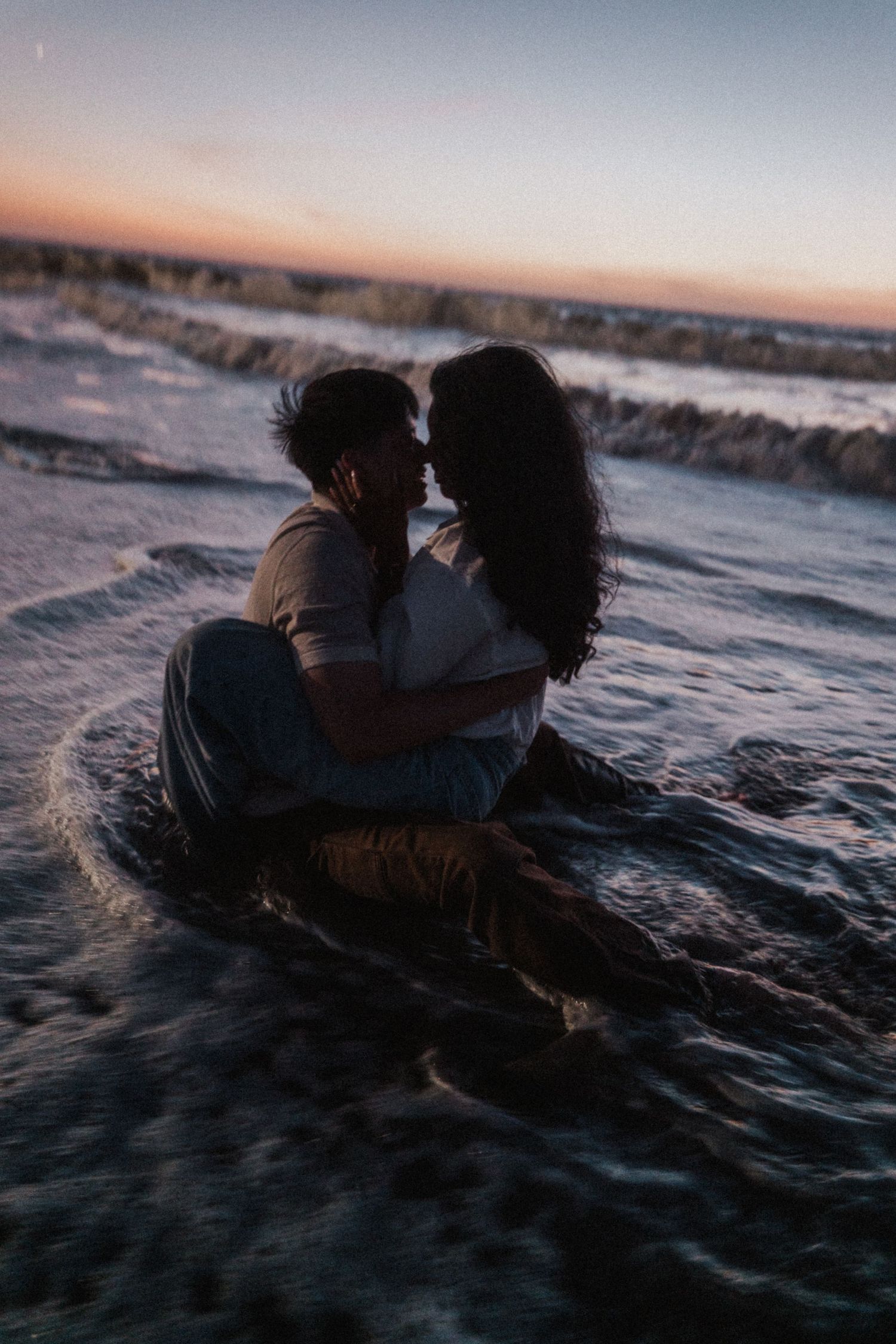 Couple sitting intimately together in shallow ocean waves during a beautiful pastel sunset on the beach.