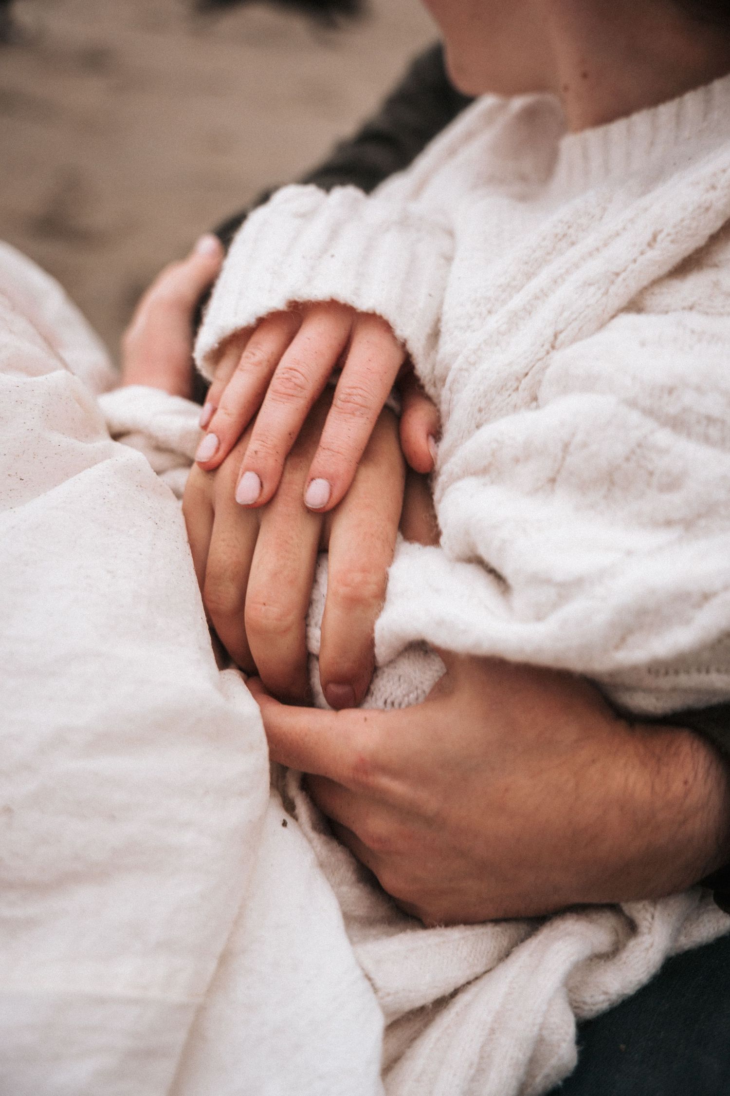 Close-up of a couple's hands tenderly intertwined, wrapped in a soft white knit blanket outdoors.
