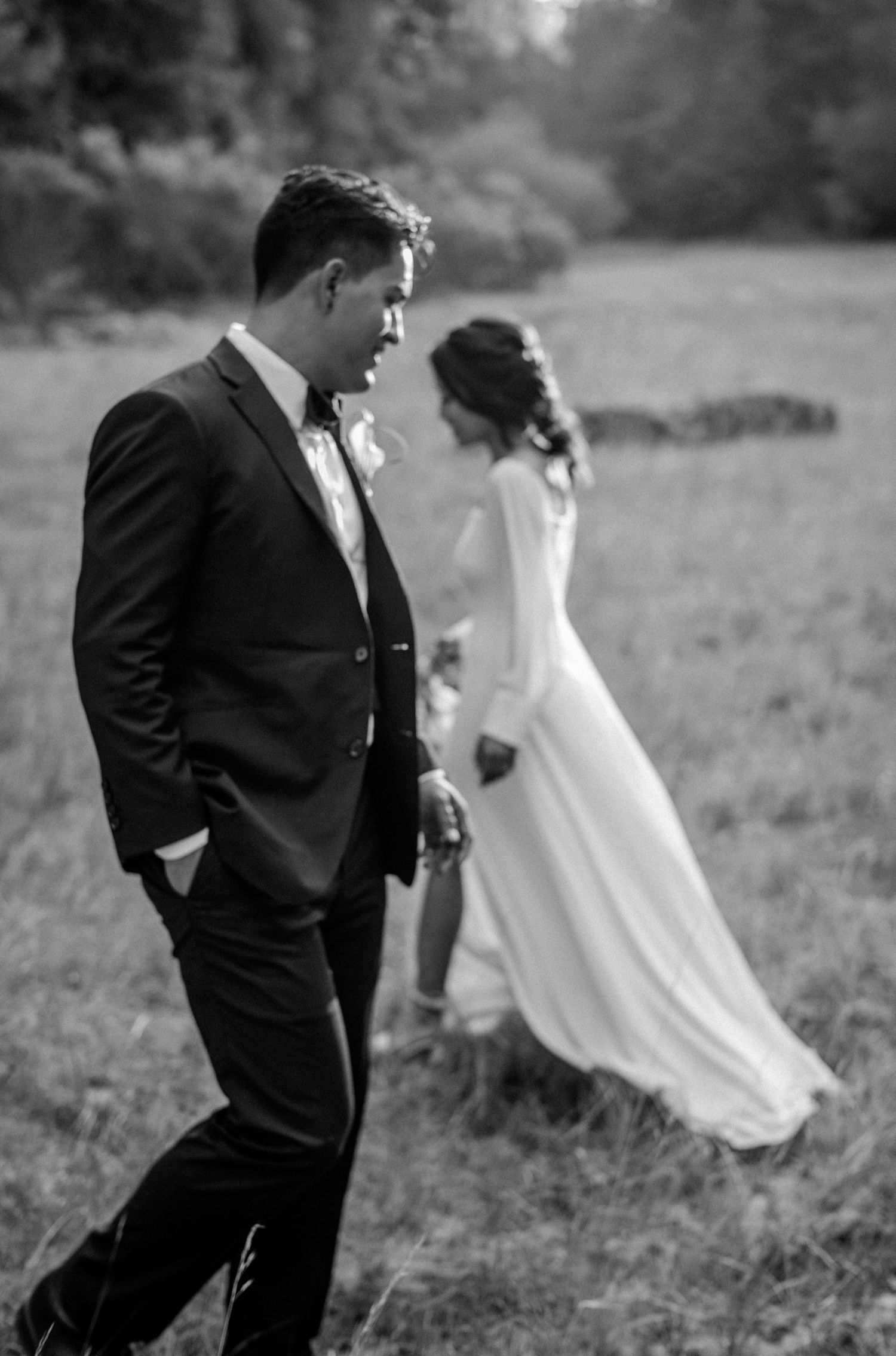 Black and white photo of a bride and groom walking through a field, holding hands in elegant wedding attire.