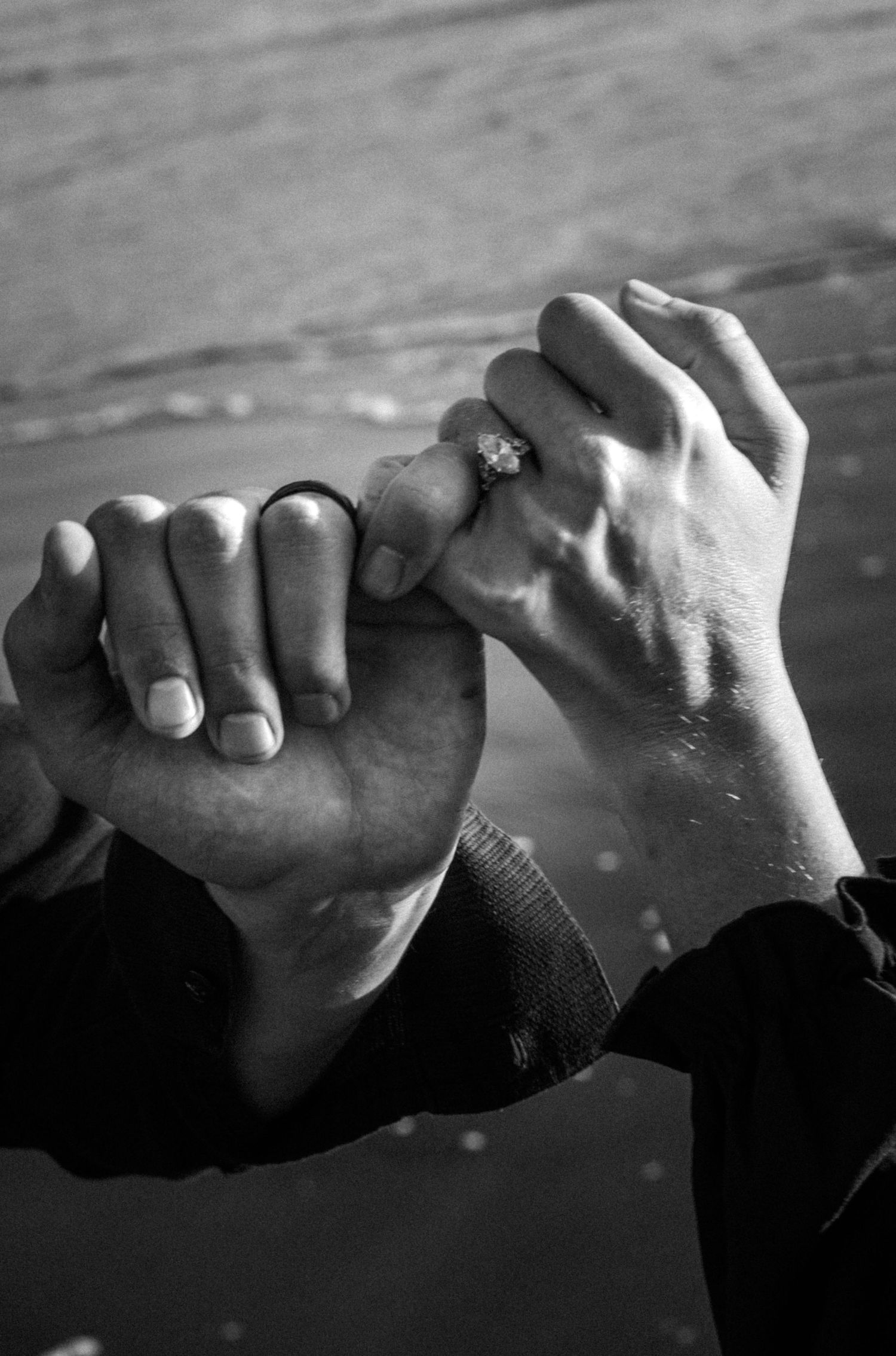 Black and white photo of two hands clasped together, one wearing an engagement ring, at a beach.