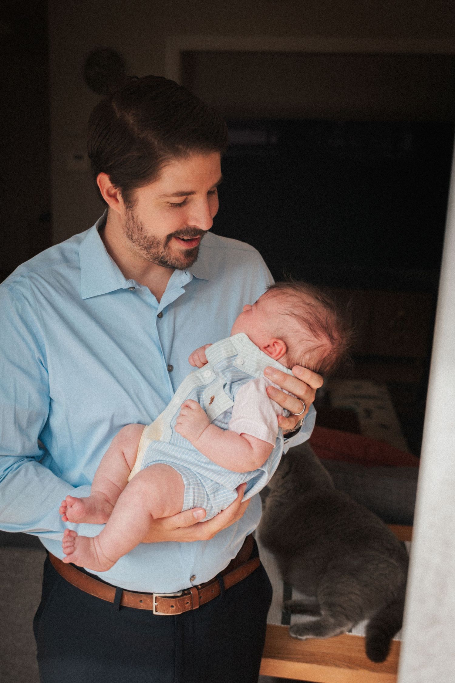 A father in a blue shirt lovingly holds his newborn baby while a gray cat looks on indoors.