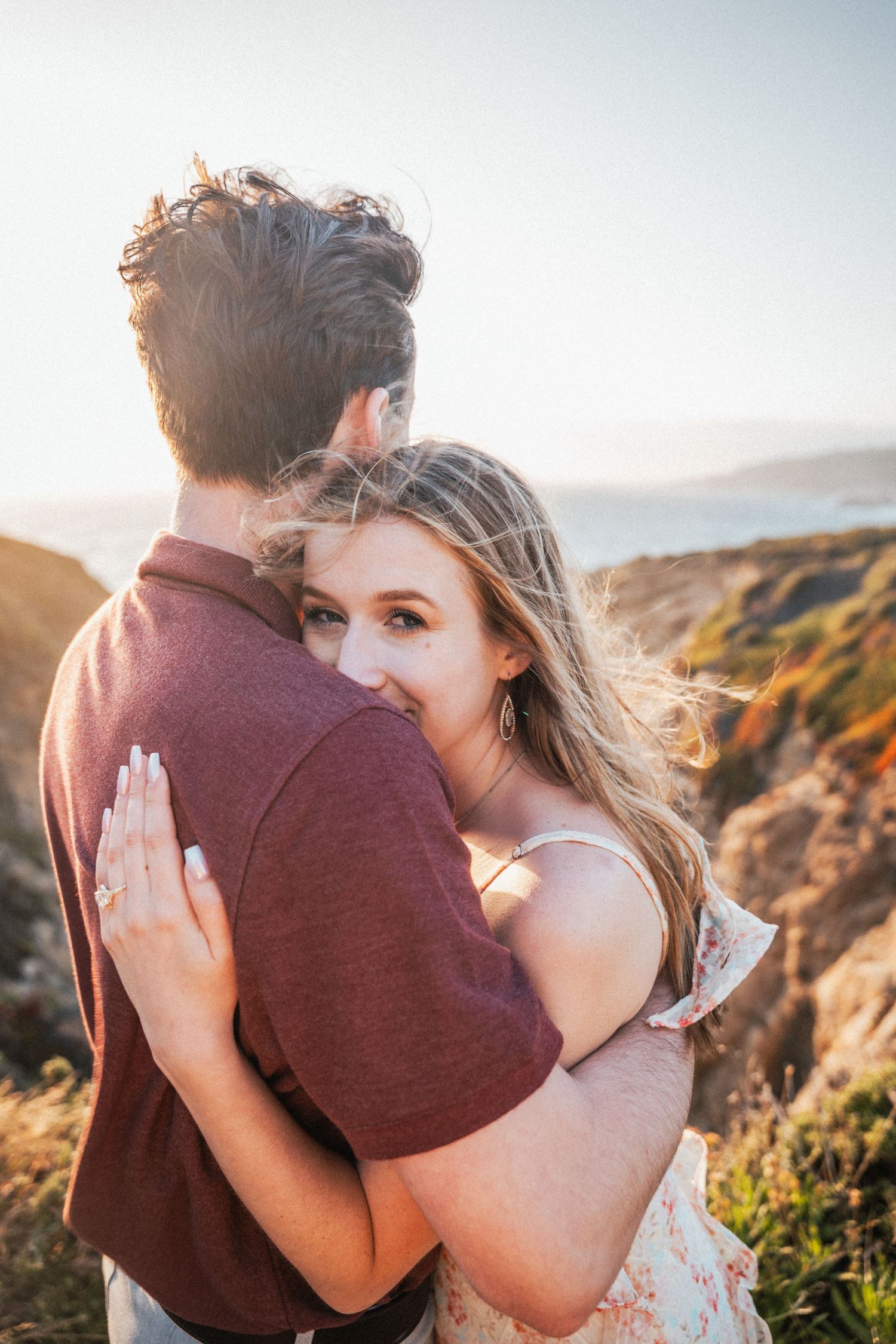 Couple embracing at golden hour on coastal cliffs, woman smiling showing engagement ring.
