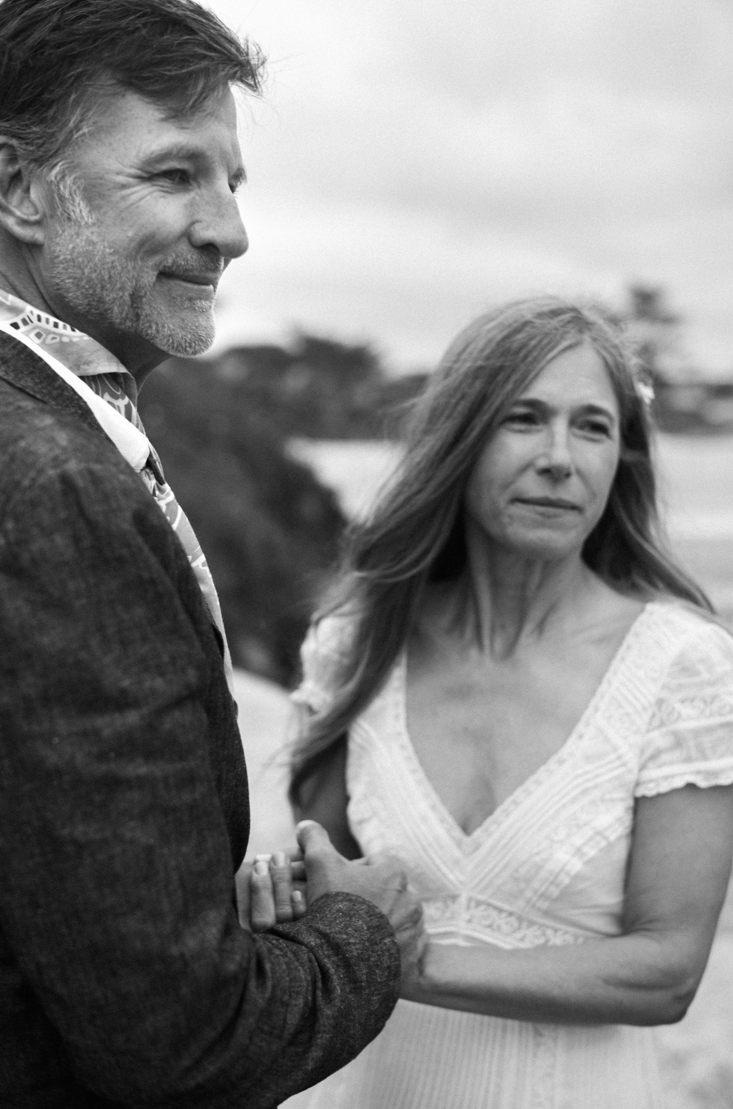 Black and white photo of a mature couple holding hands on a beach, woman in white lace dress, man in gray jacket.