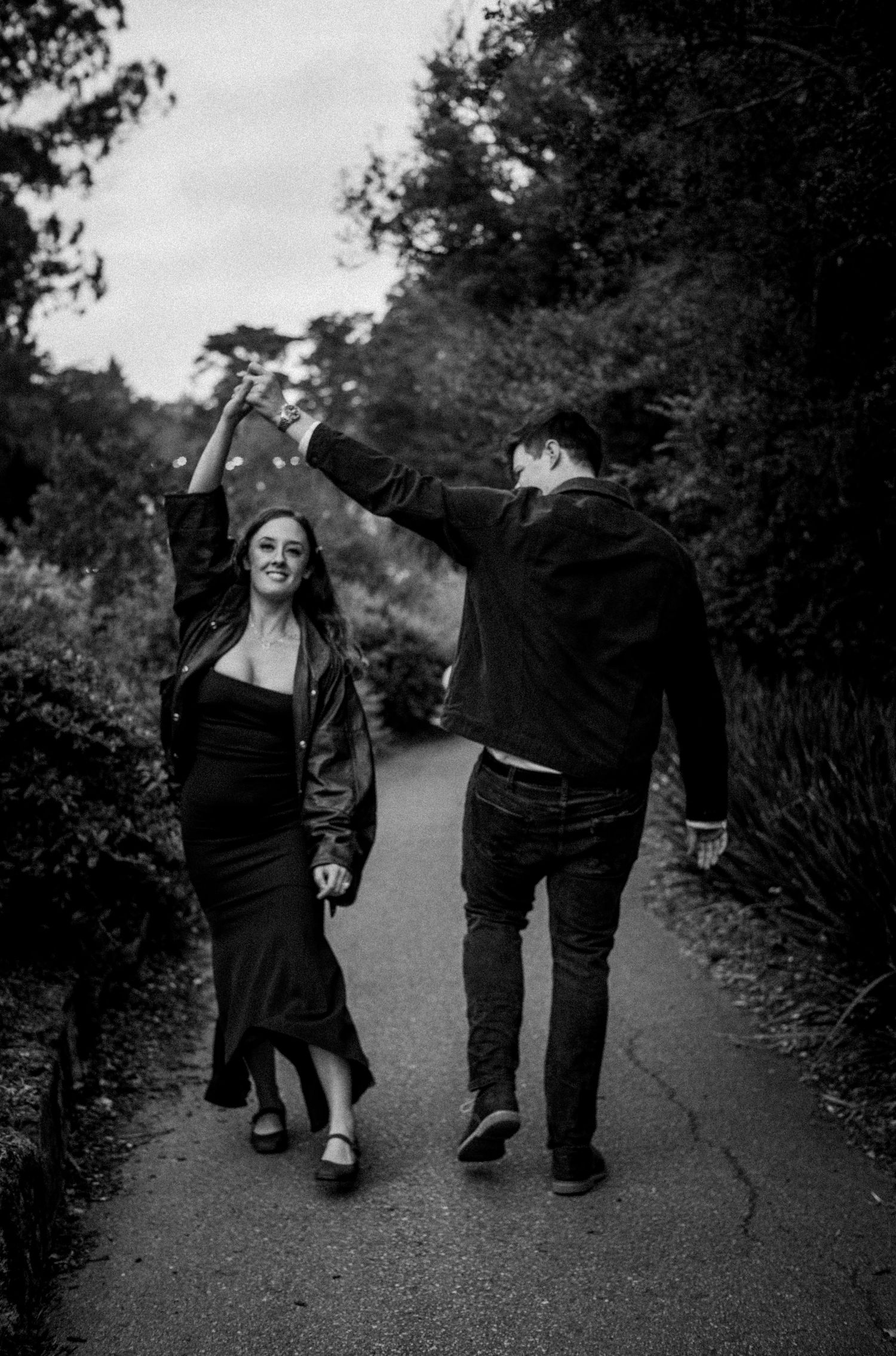 Black and white photo of couple dancing on a tree-lined park pathway.