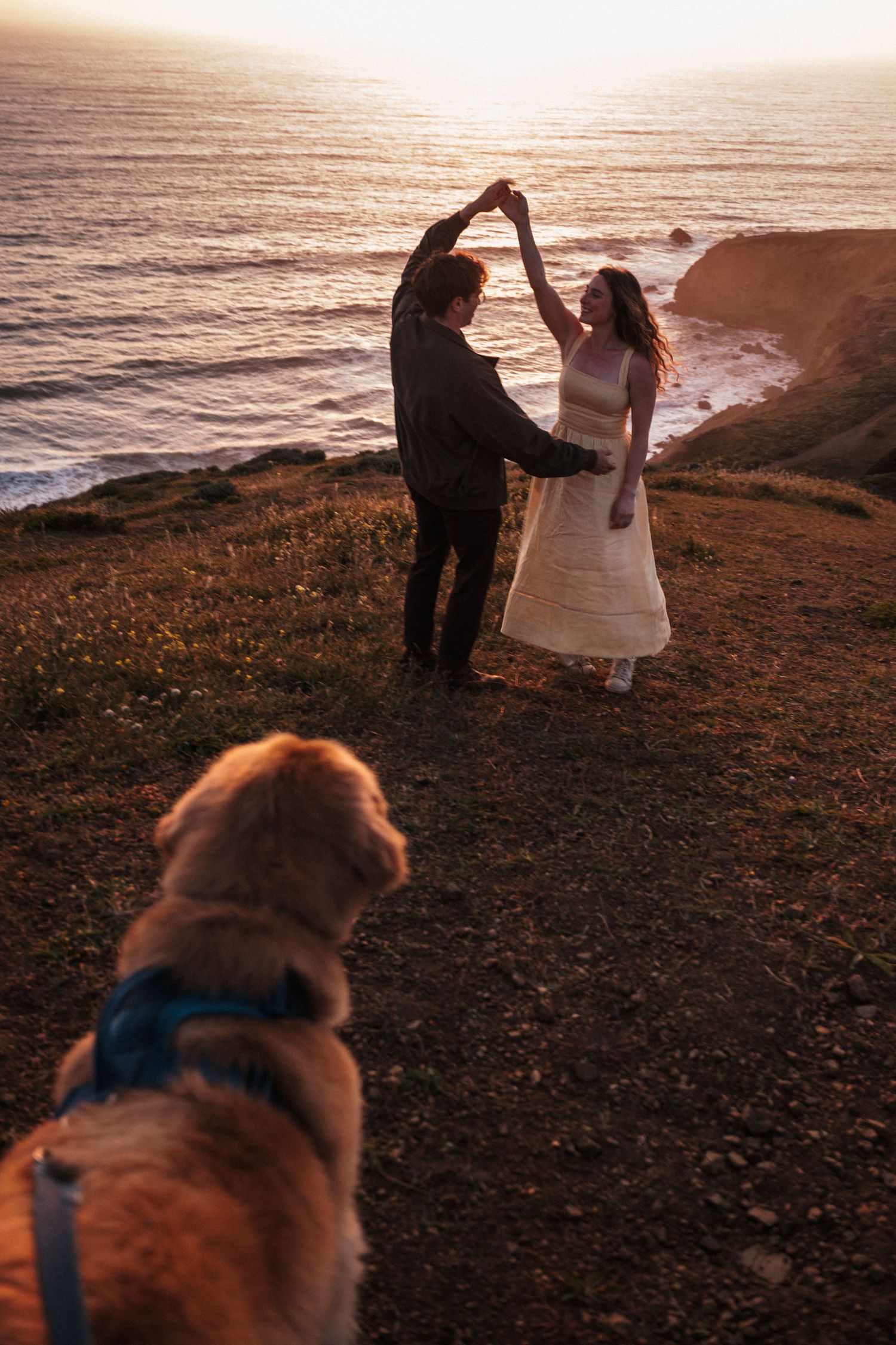 Couple dancing on coastal cliffside at sunset with golden retriever dog in foreground and ocean waves below.