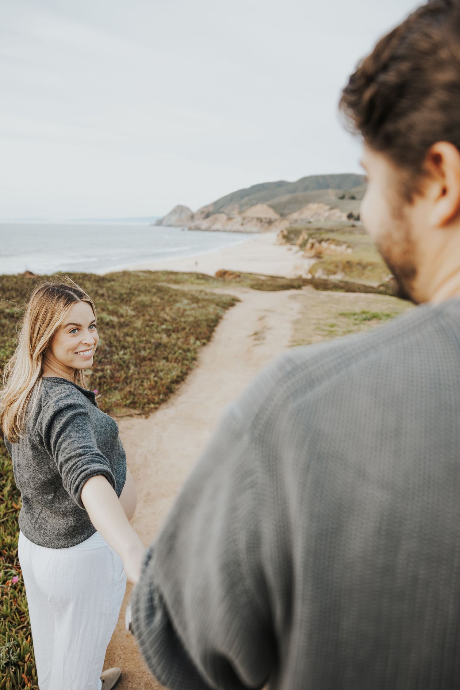 Smiling blonde woman holds mans hand on scenic coastal cliffside path with ocean views in background.