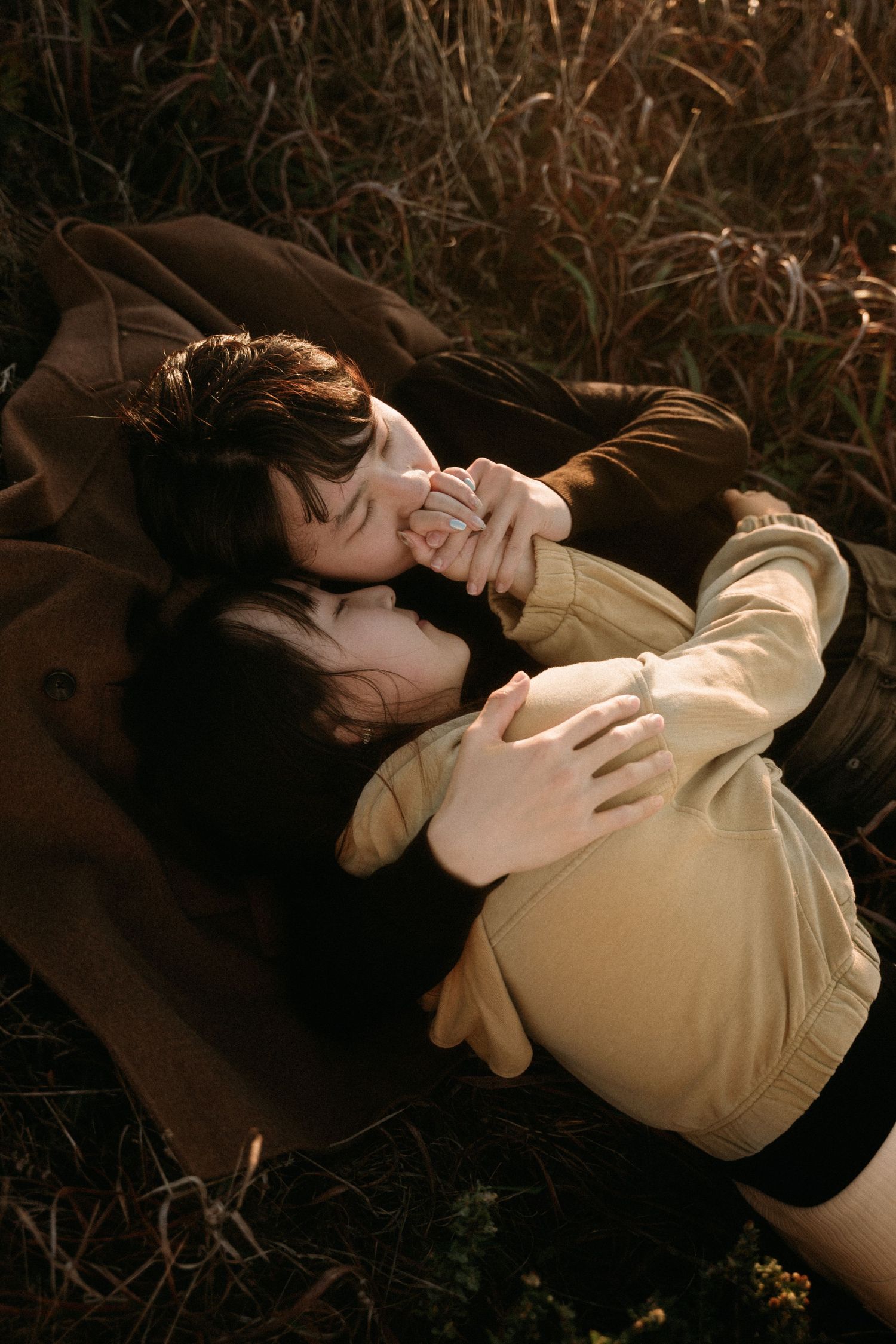 Two people in warm-toned jackets lie close together in dry grass at golden hour, sharing an intimate, tender embrace.