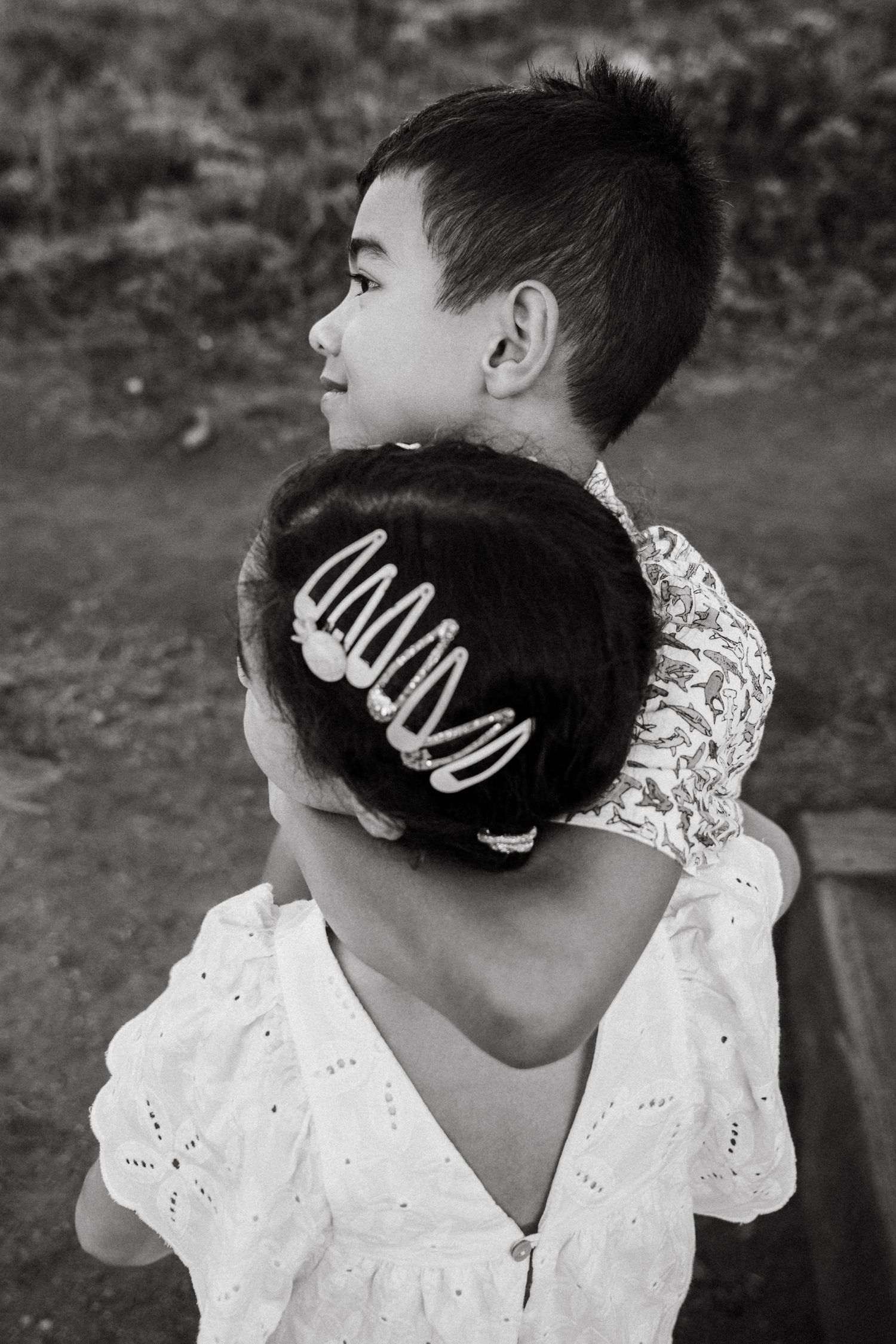 Black and white photo of two children hugging, girl with hair clips in bun, boy looking away outdoors.
