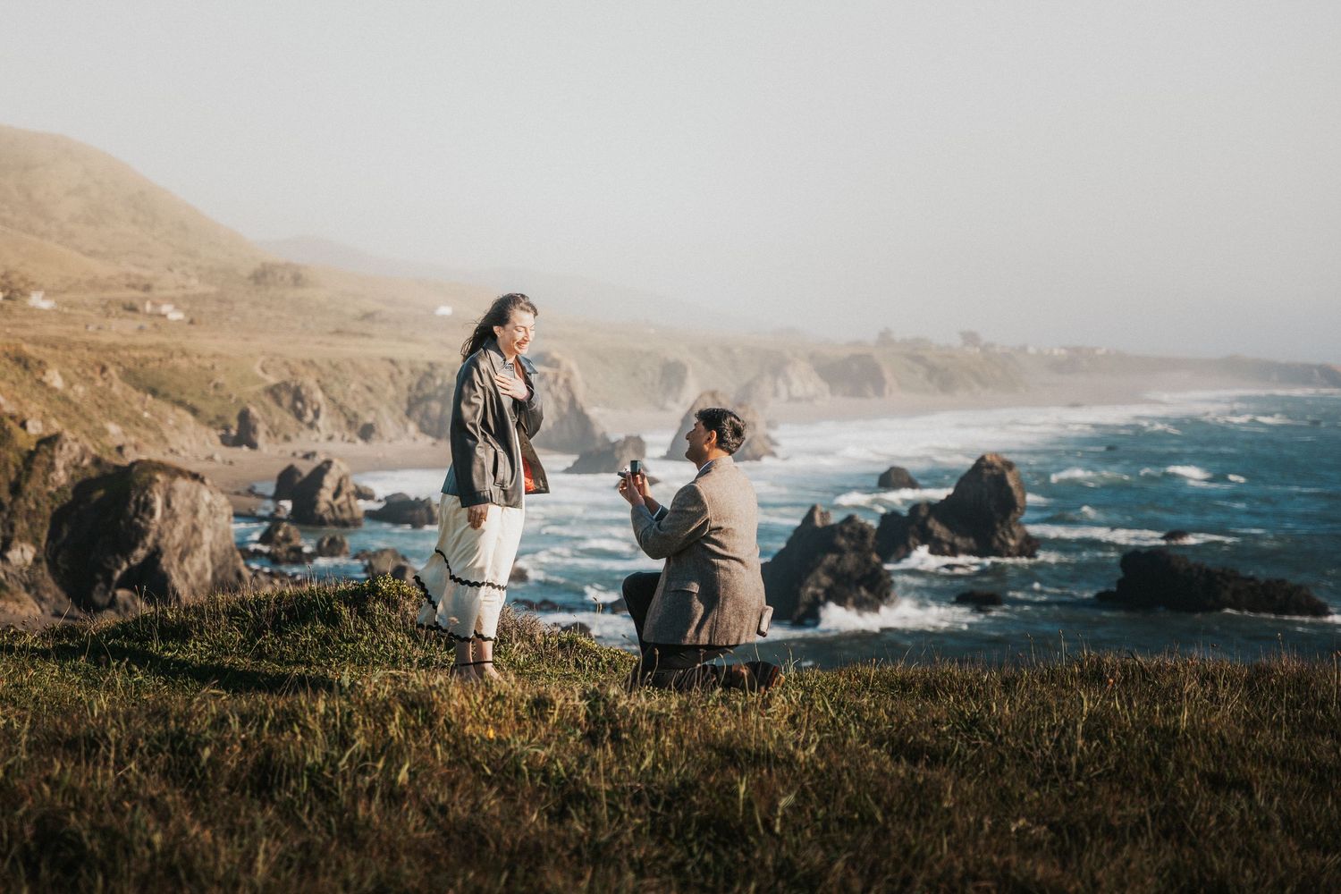 Man proposing to woman on a scenic coastal cliff with rocky shoreline and ocean waves in the background.