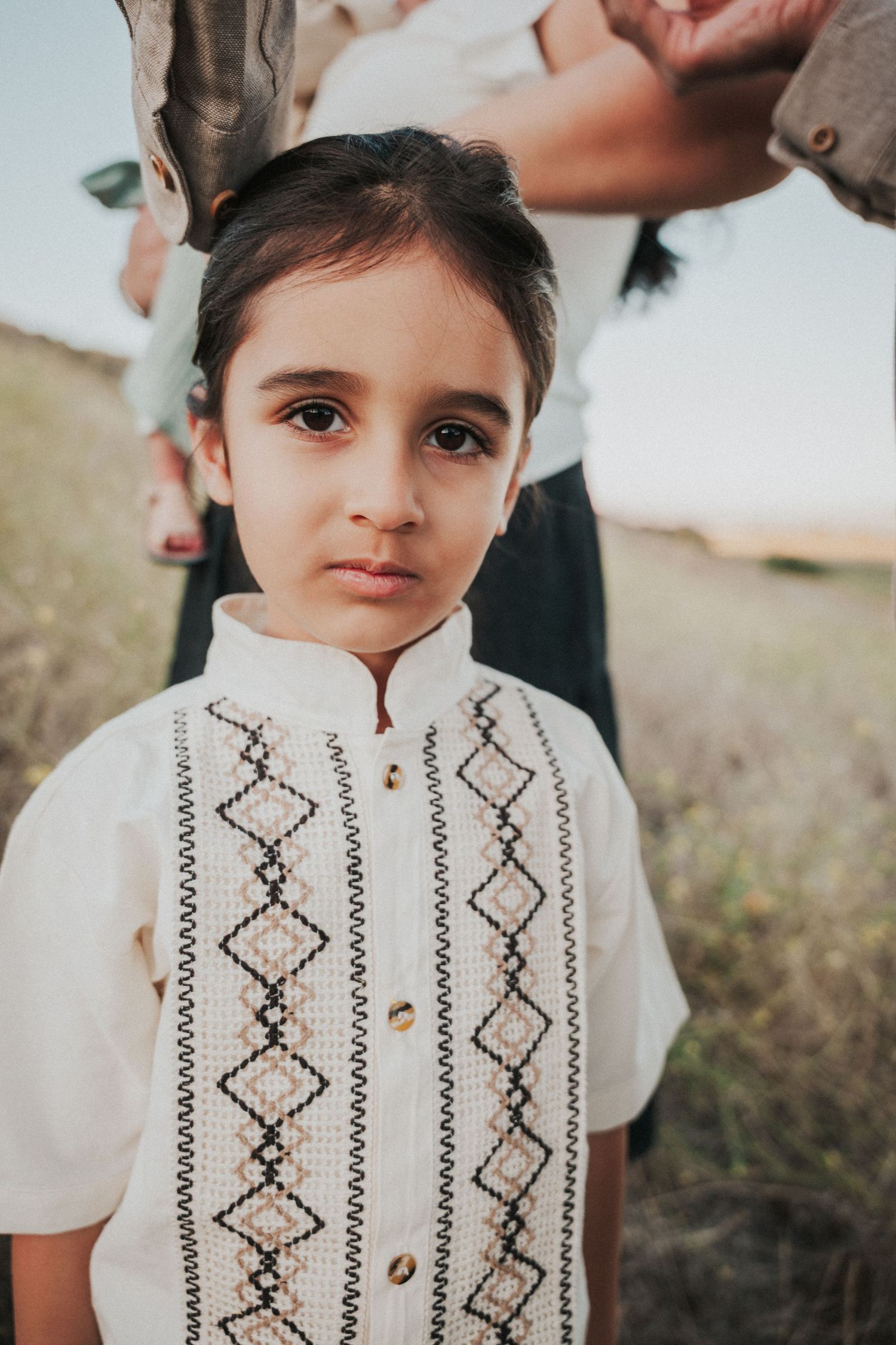 Young boy in embroidered white traditional shirt gazes at camera in a field.