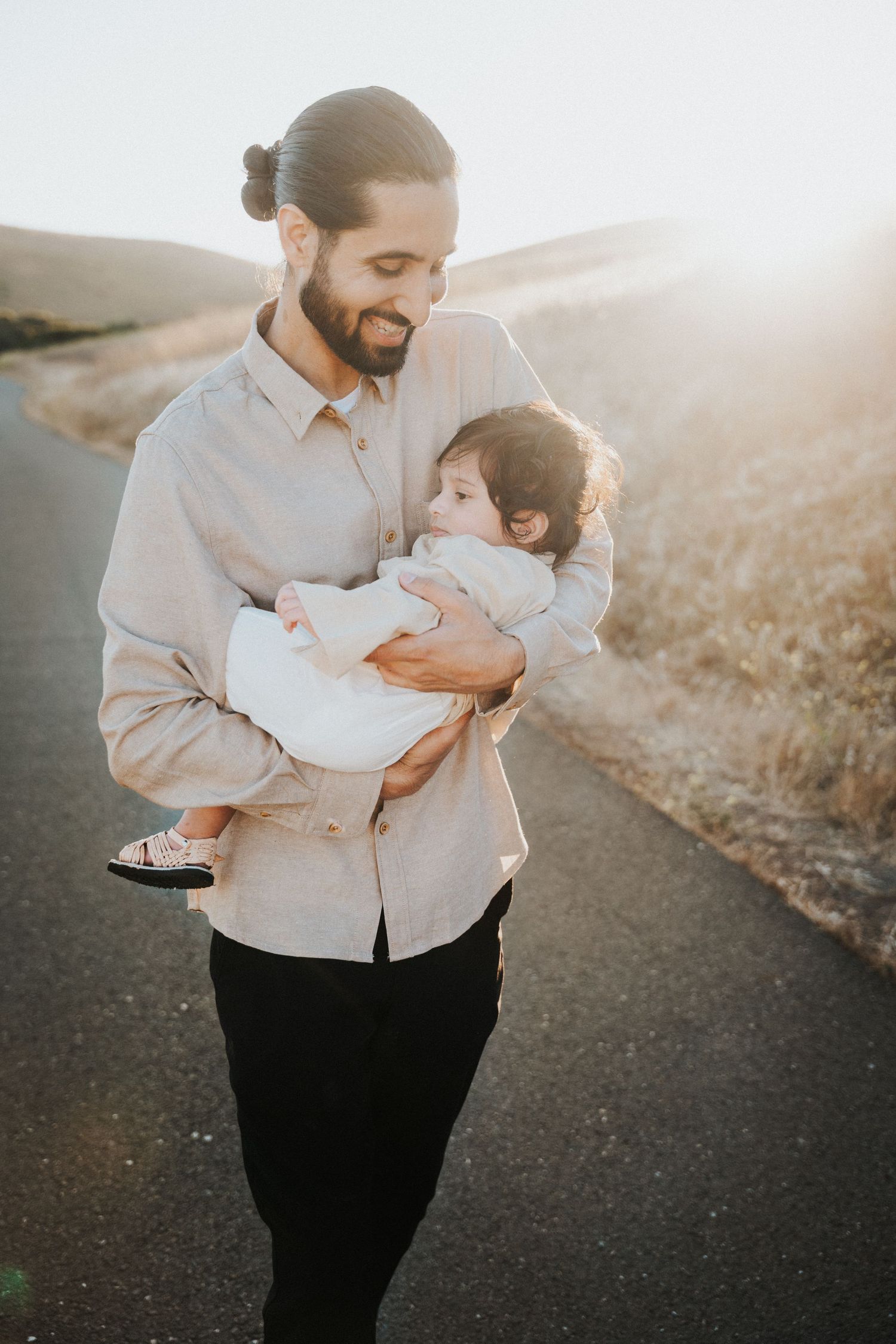 Smiling bearded father with man bun holding baby outdoors at golden hour on a sunlit road.