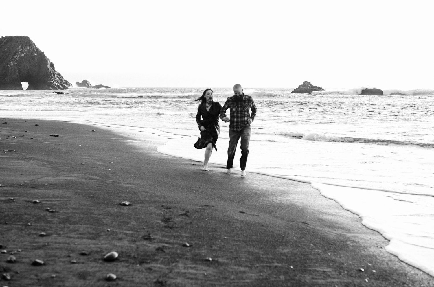 Black and white photo of a couple joyfully running together along a scenic beach with rocky outcroppings in the background.